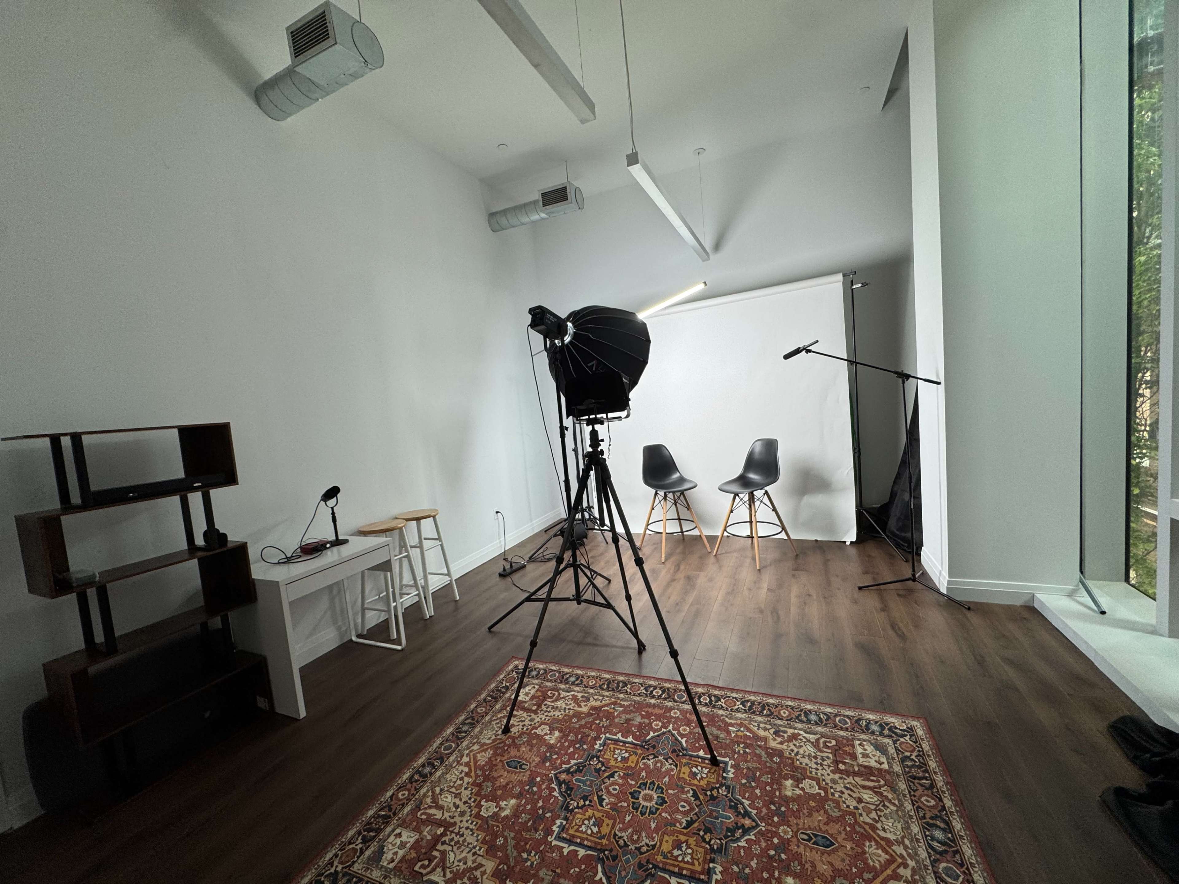 The image shows a bright, empty studio space featuring two chairs, a backdrop, and lighting equipment, with a wooden floor and a large window.