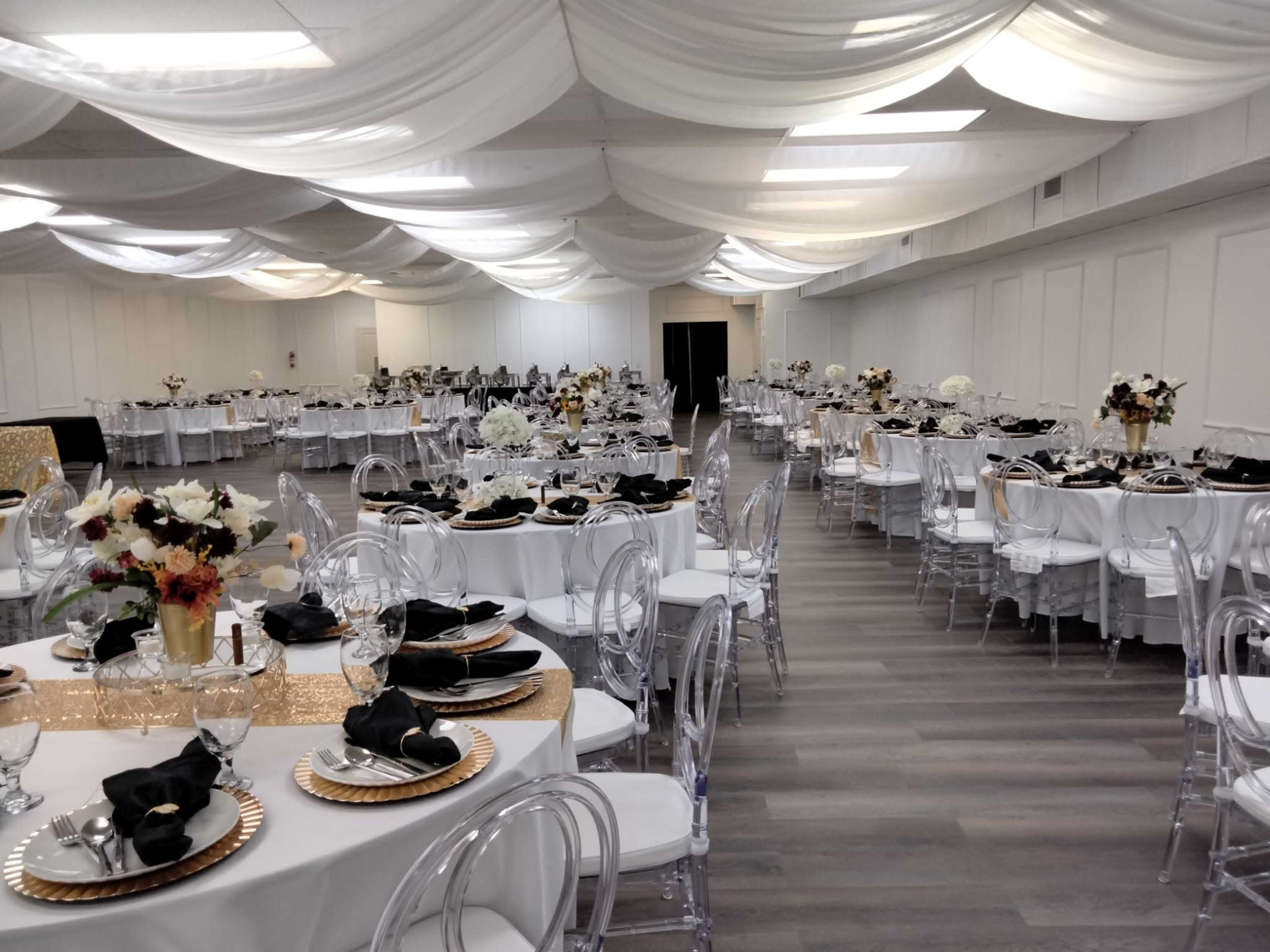 The image shows a large banquet hall filled with elegantly set tables, each adorned with white tablecloths, black napkins, and decorative centerpieces.