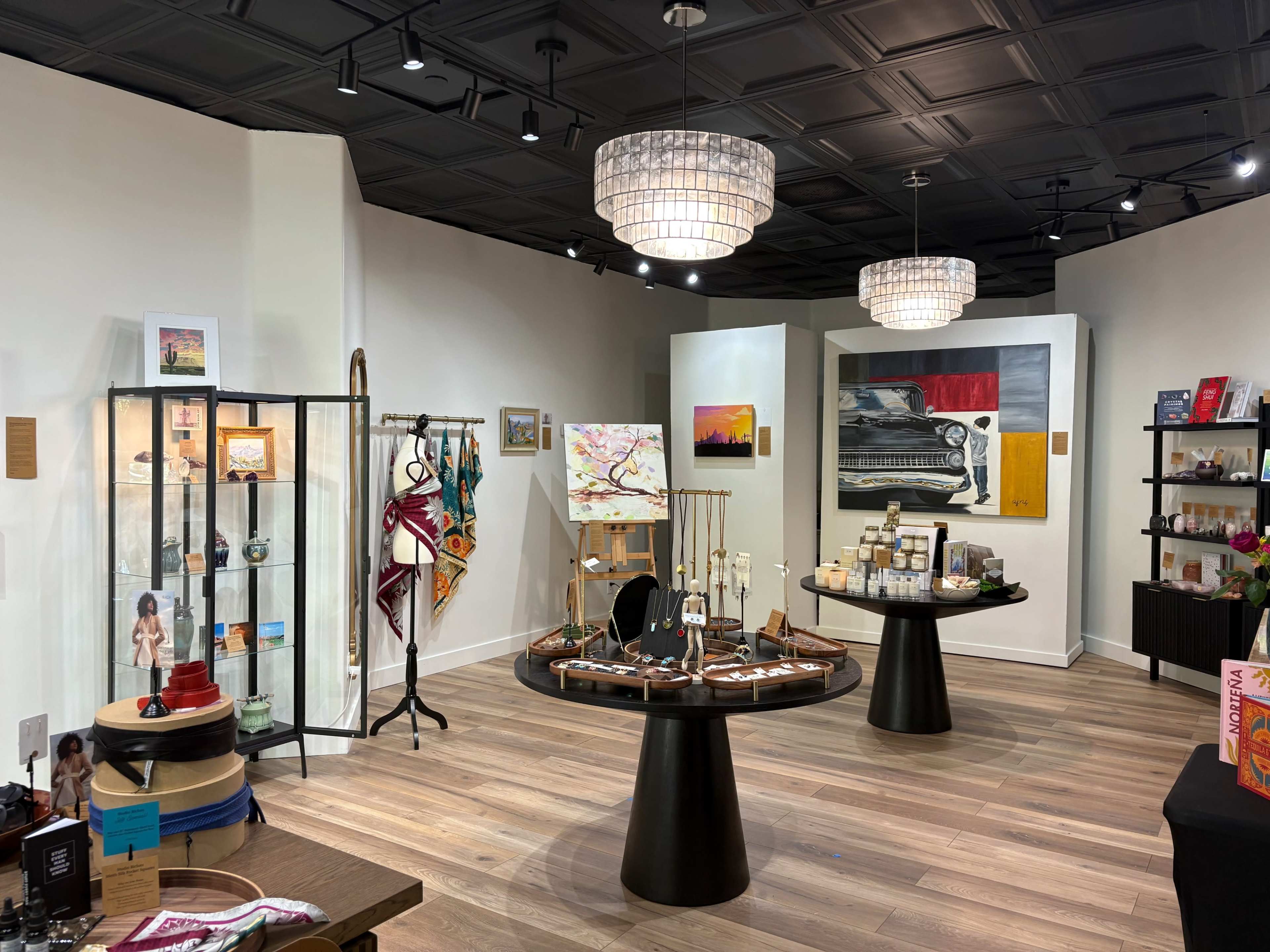 The image shows a contemporary retail store interior featuring various display tables, artwork, and decorative items under modern lighting.