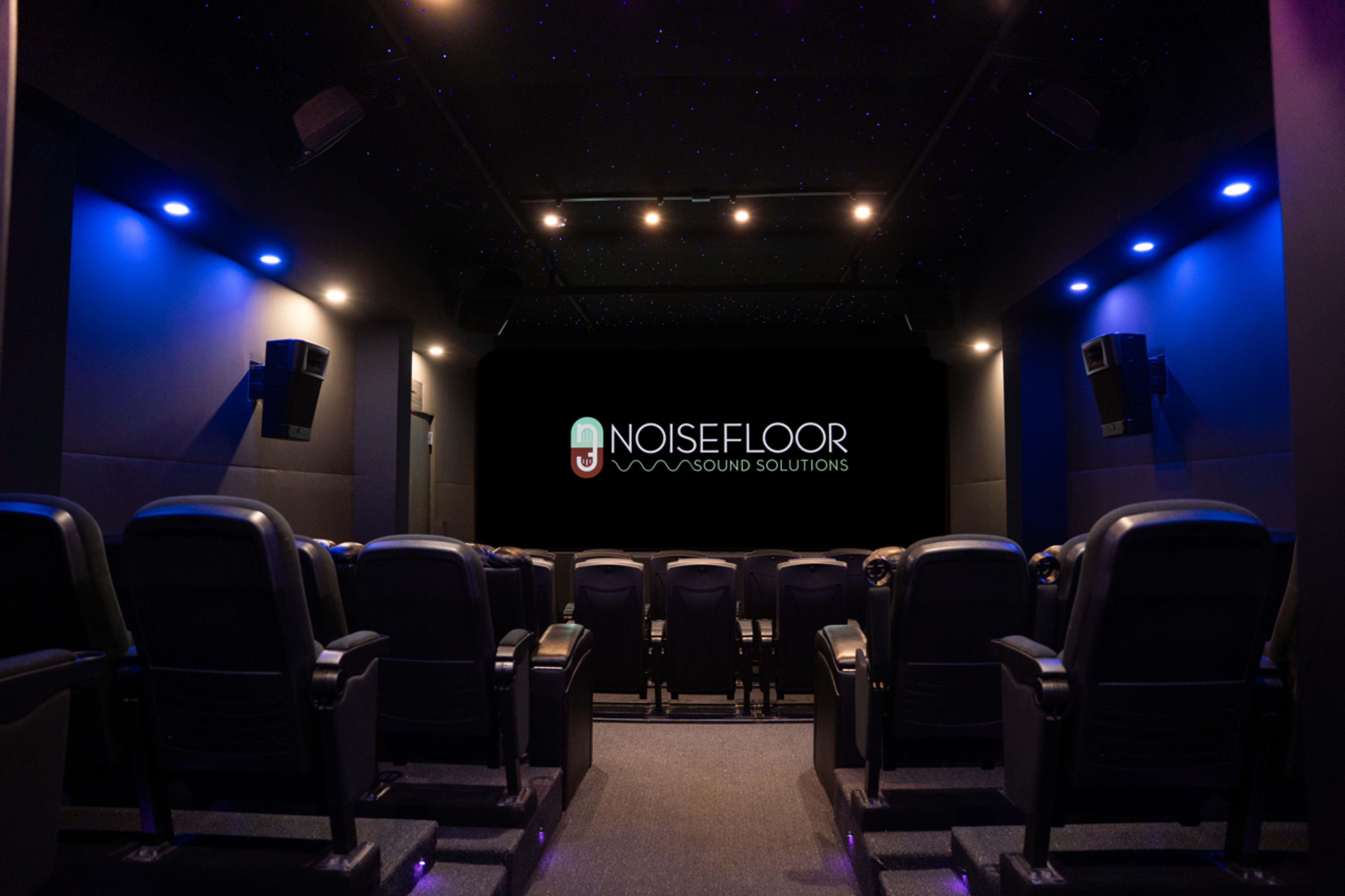 The image shows a dark theater interior with rows of black seating facing a large screen featuring the logo for "NoiseFloor Sound Solutions."