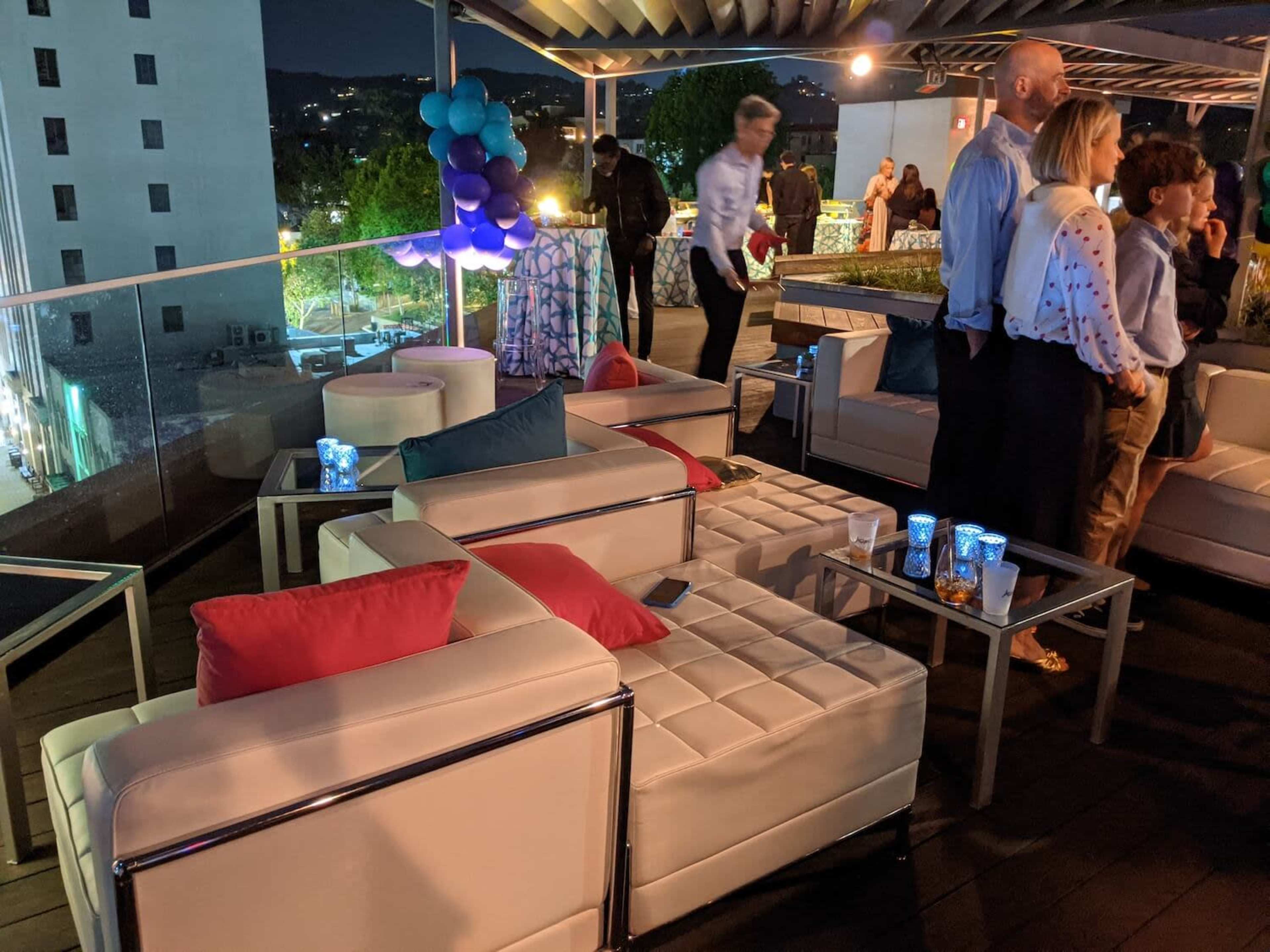 The image shows a rooftop terrace at night, featuring modern white furniture with colorful cushions, people mingling, and city lights in the background.