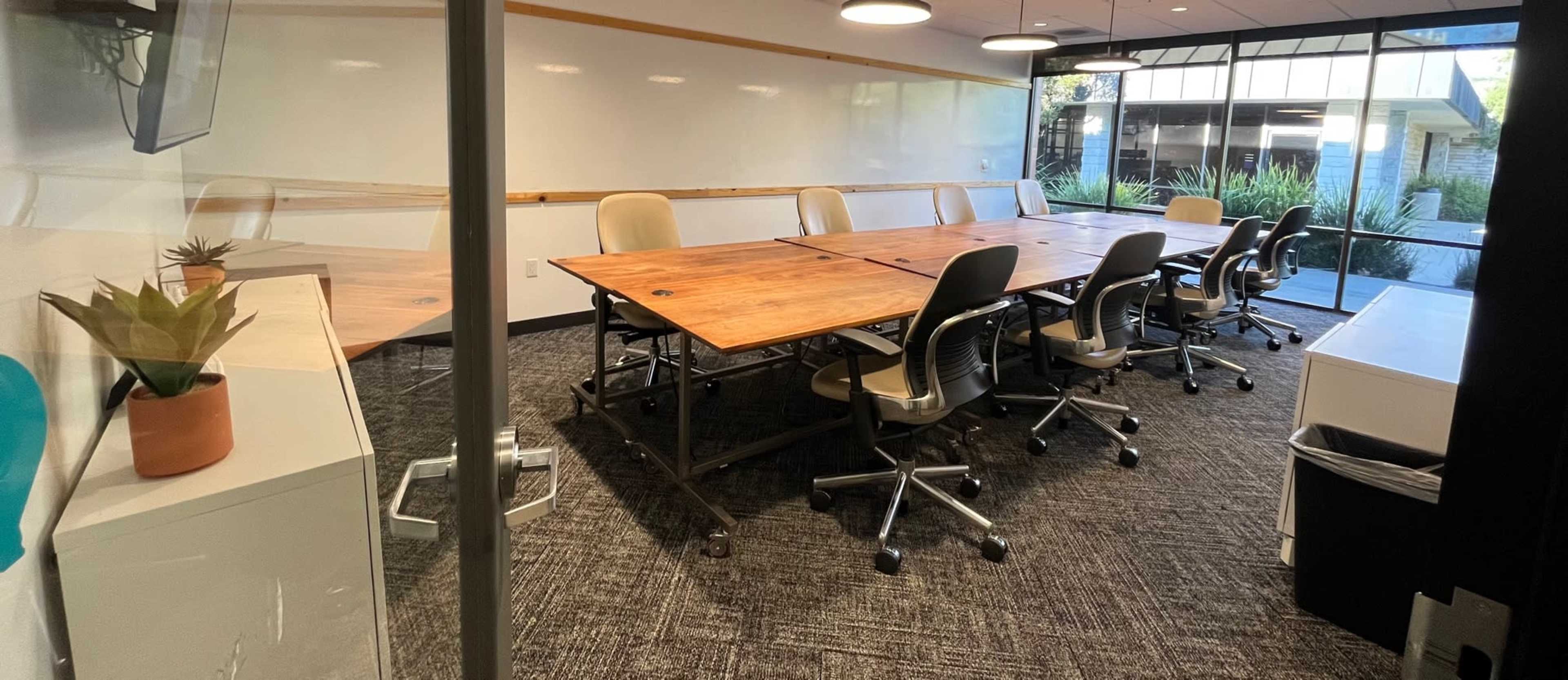 A modern conference room with a large wooden table surrounded by ergonomic chairs and large windows allowing natural light.