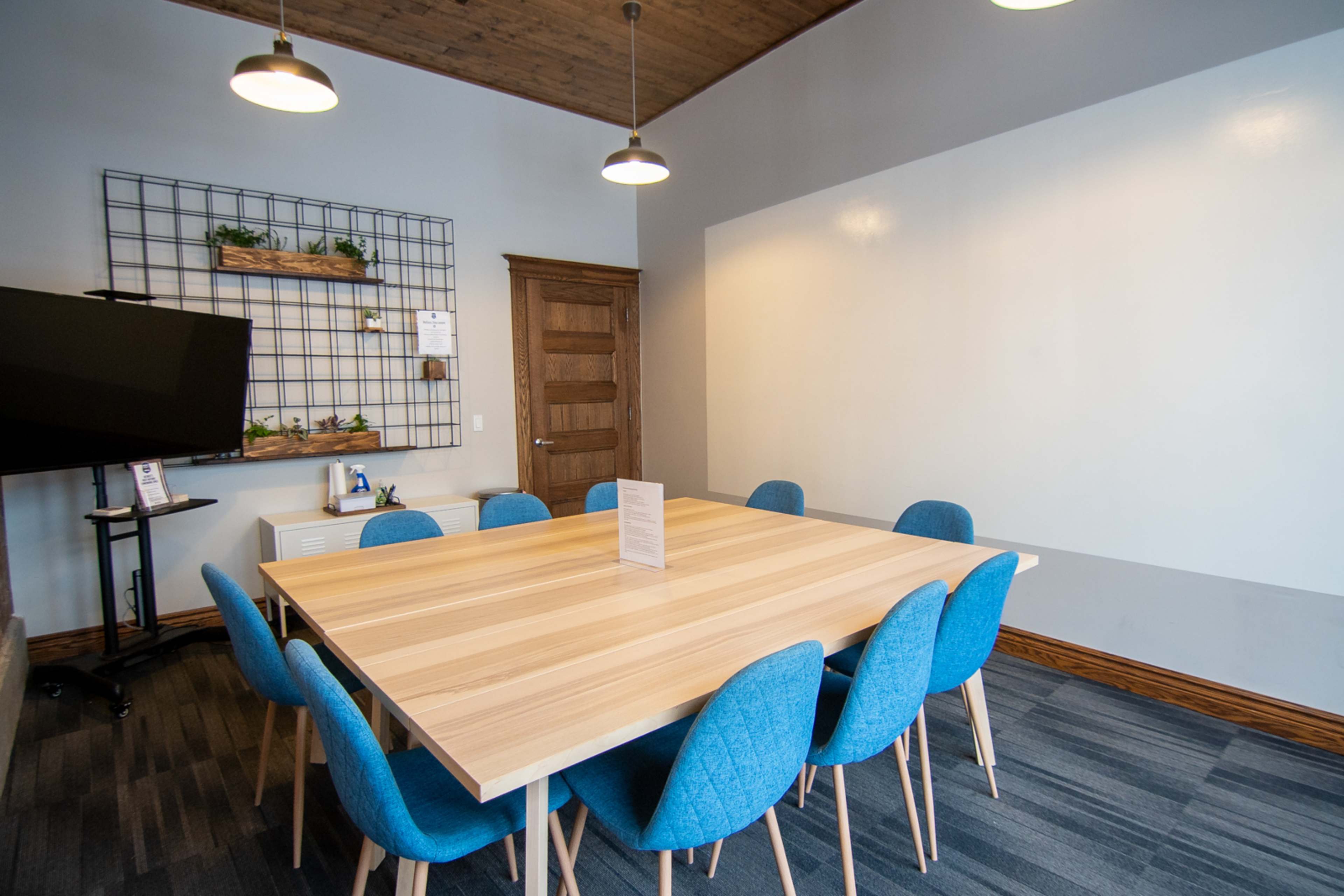 A meeting room features a large wooden table surrounded by eight blue chairs, with a wall-mounted shelf and a TV in the background.