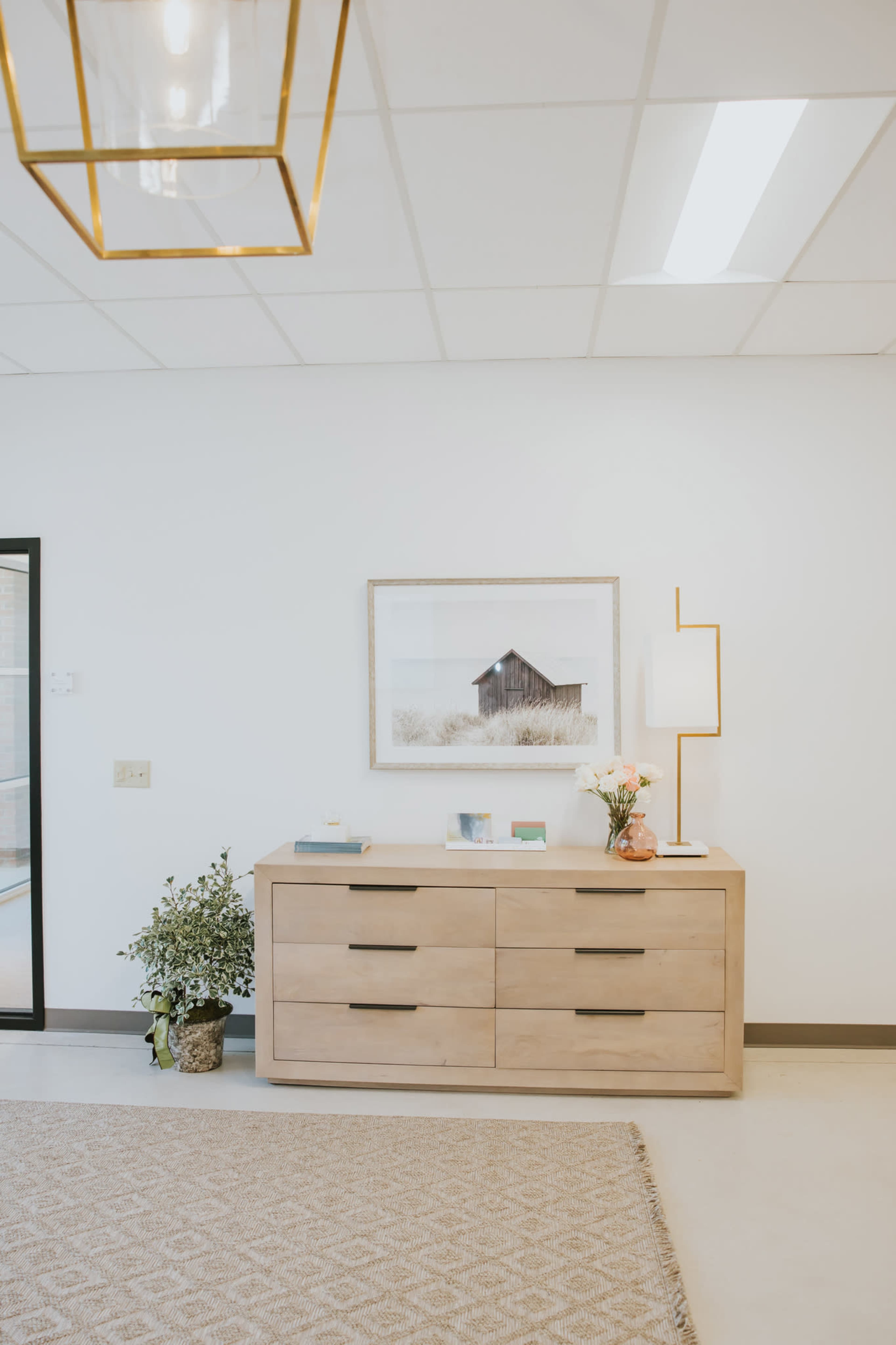 The image shows a light-filled room featuring a wooden dresser with six drawers, a framed artwork on the wall above it, and a floor lamp beside a floral arrangement.