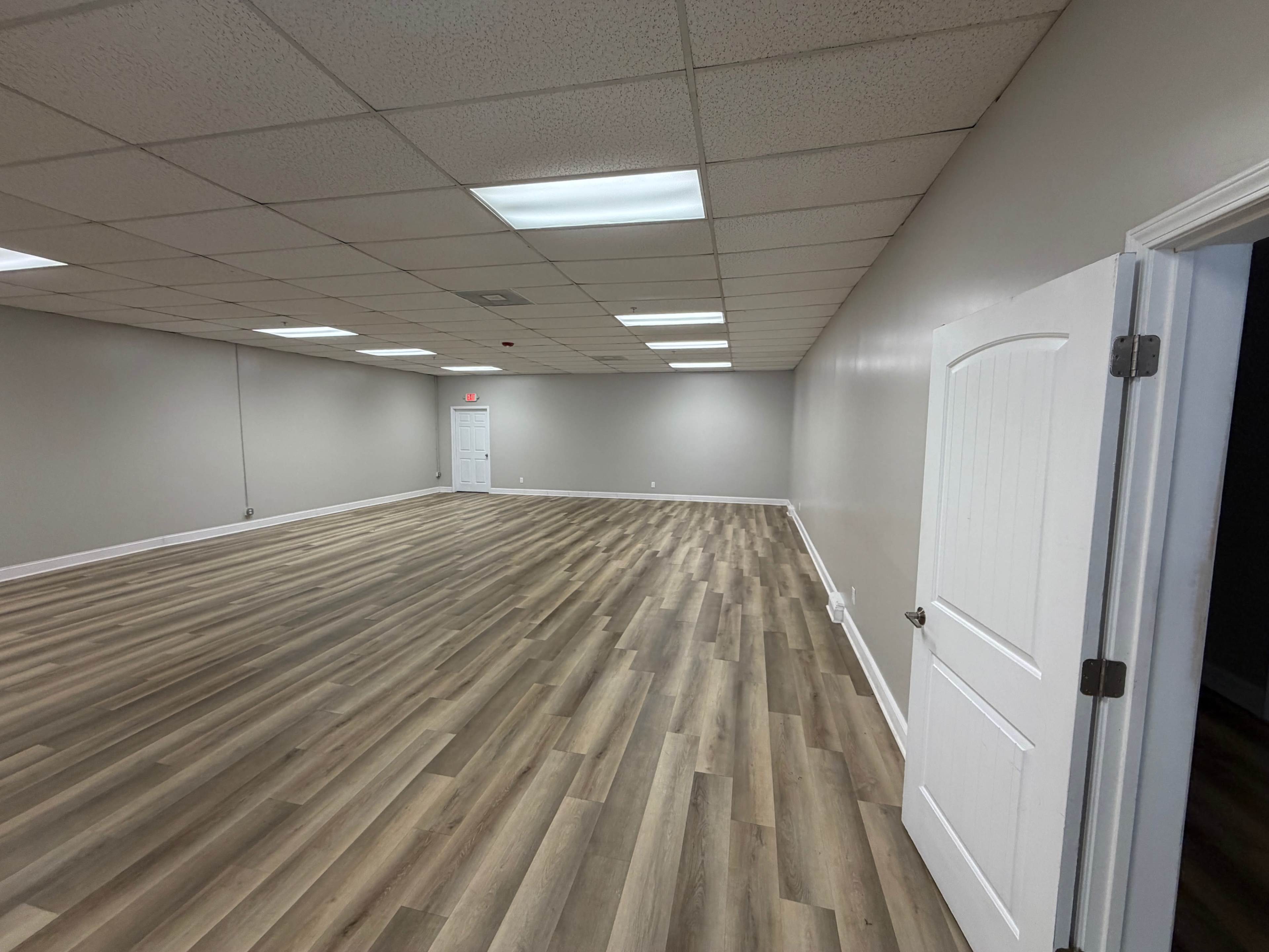 The image shows a spacious, empty room with wooden flooring and gray walls, illuminated by overhead fluorescent lights.