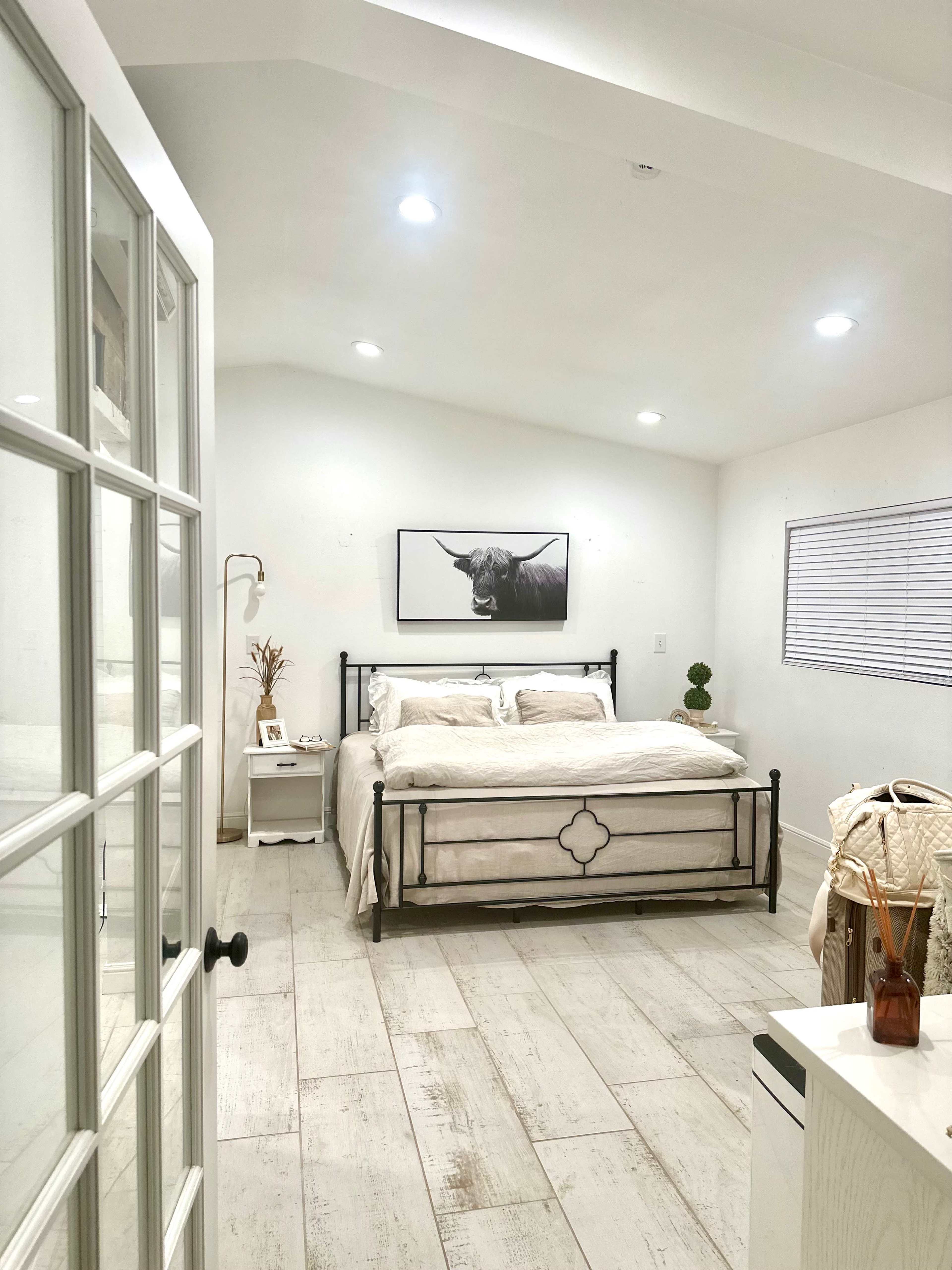 A bright bedroom features a metal bed frame with white bedding, a black-and-white bull print on the wall, and large windows with white blinds.