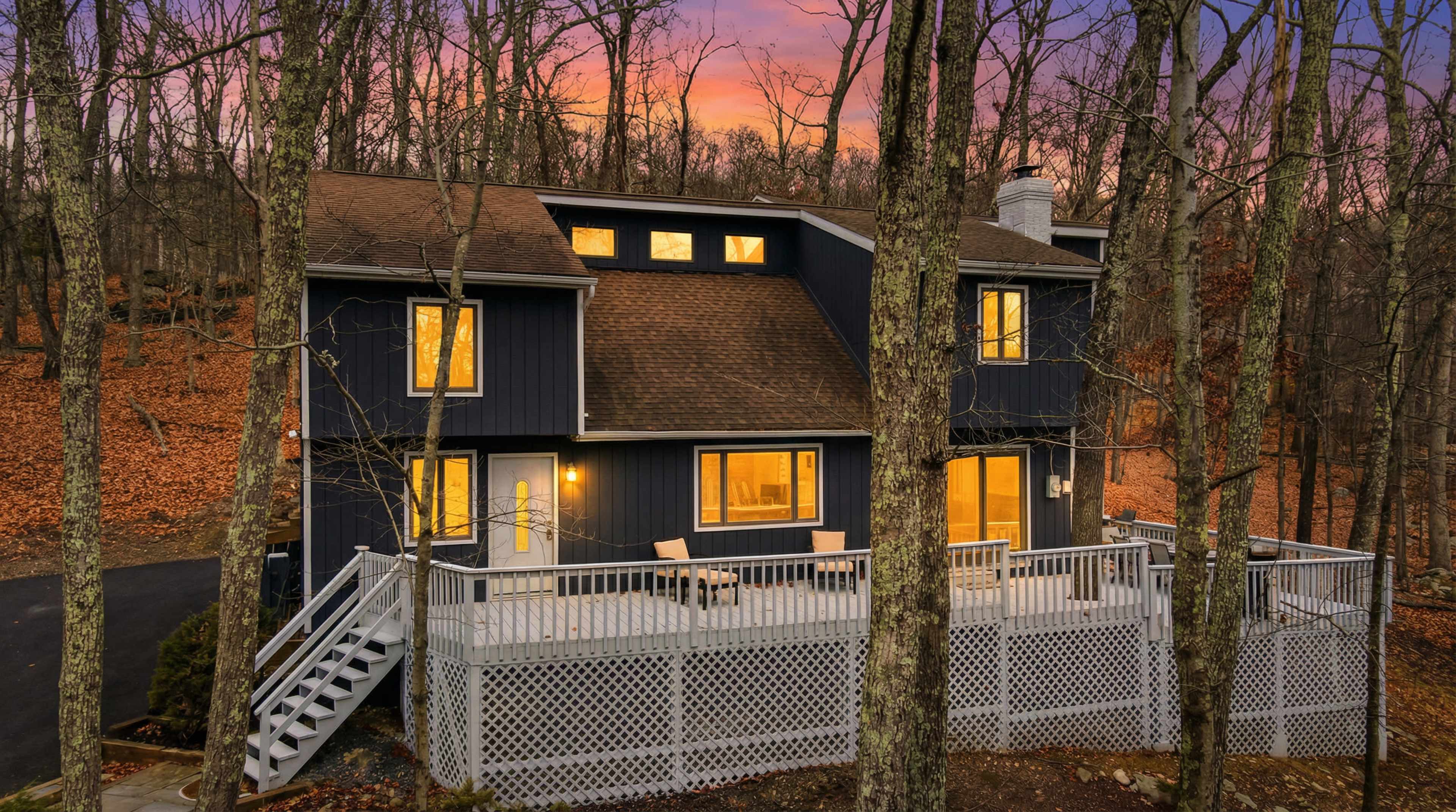 A modern dark-colored house with large windows and a deck is surrounded by trees and autumn foliage at dusk.