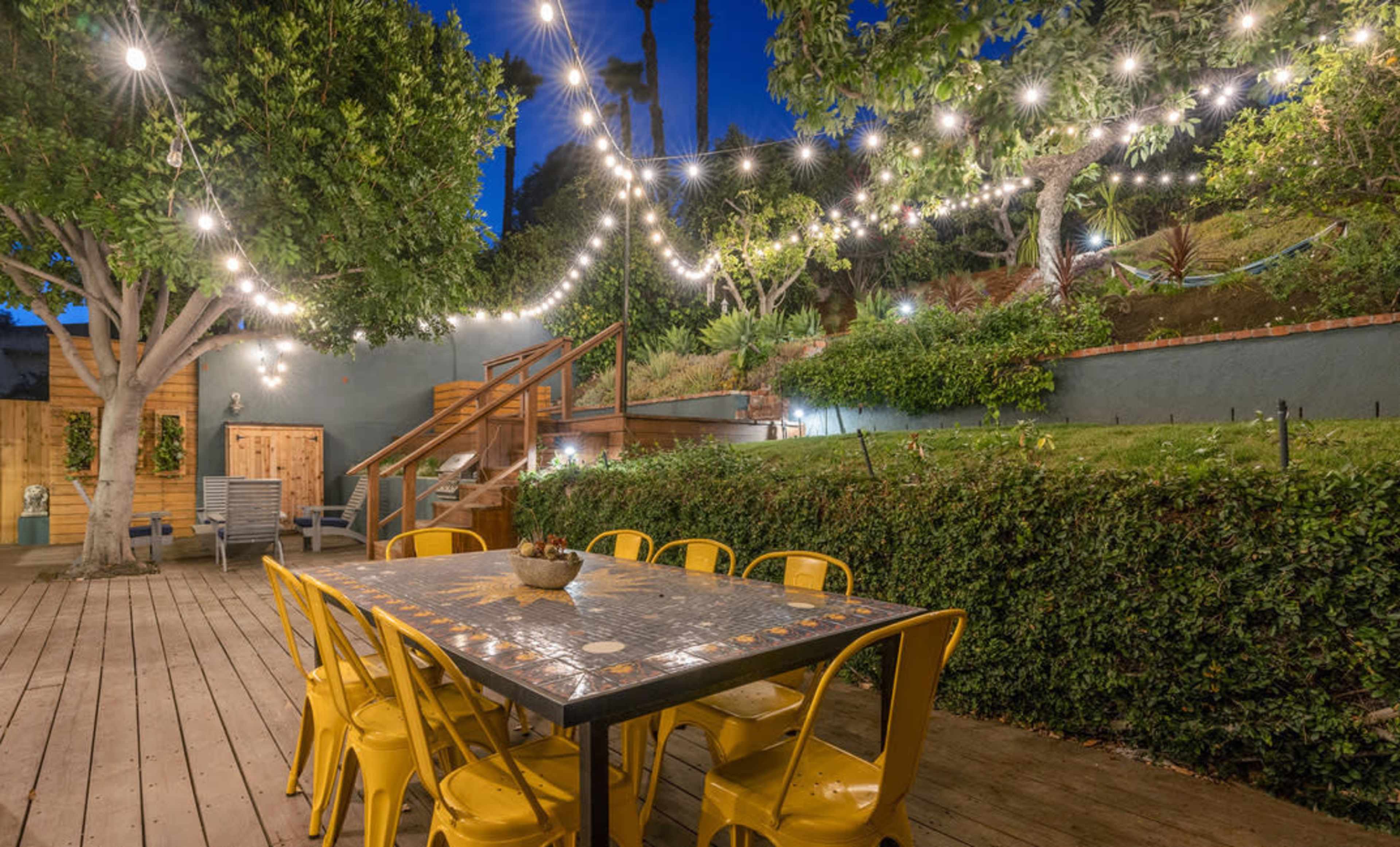 A wooden deck features a large table surrounded by yellow chairs, illuminated by string lights, with a landscaped area and stairs in the background.