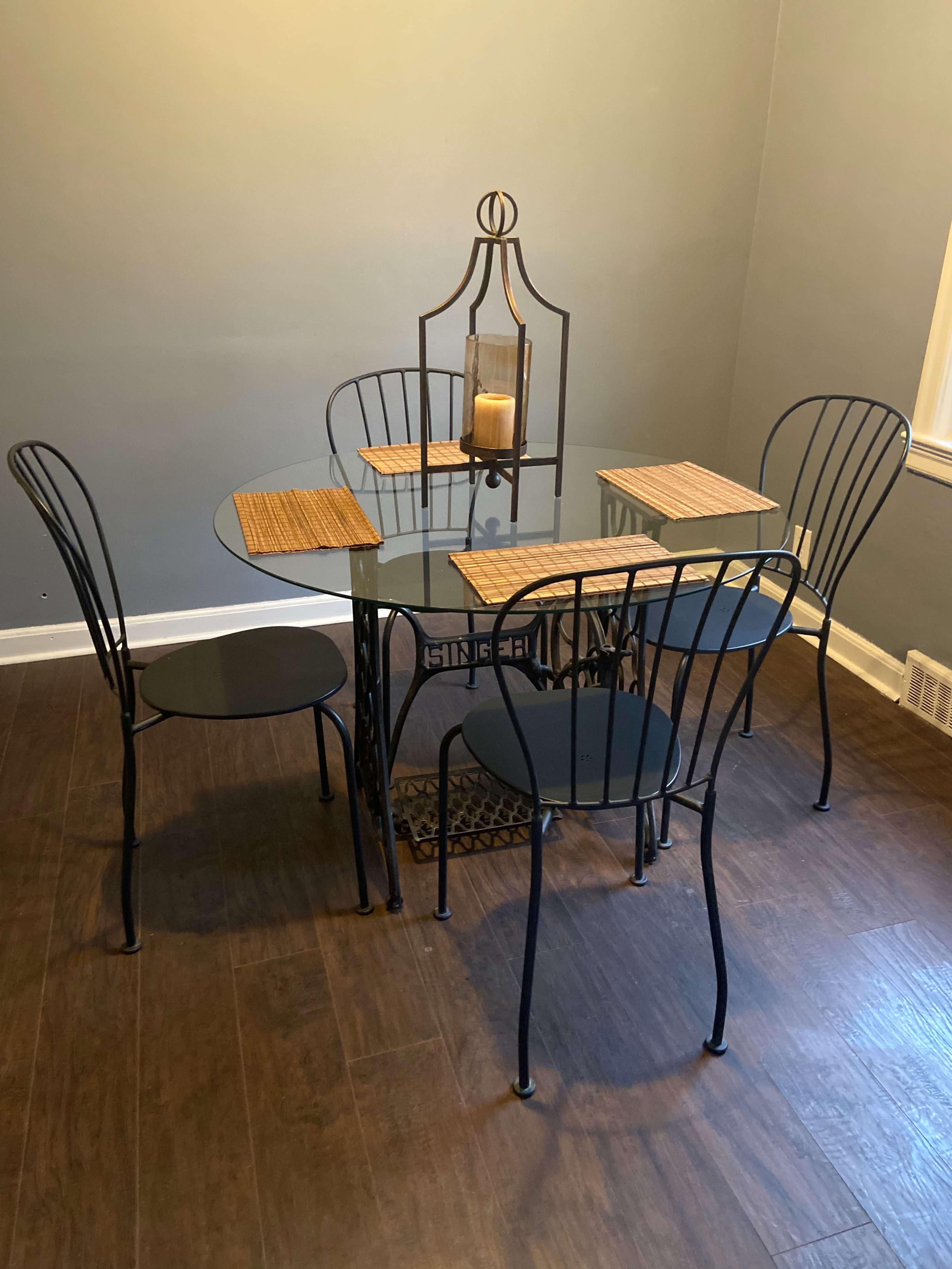 A glass-topped dining table is set with four black metal chairs and two placemats, with a candle in a lantern centerpiece.