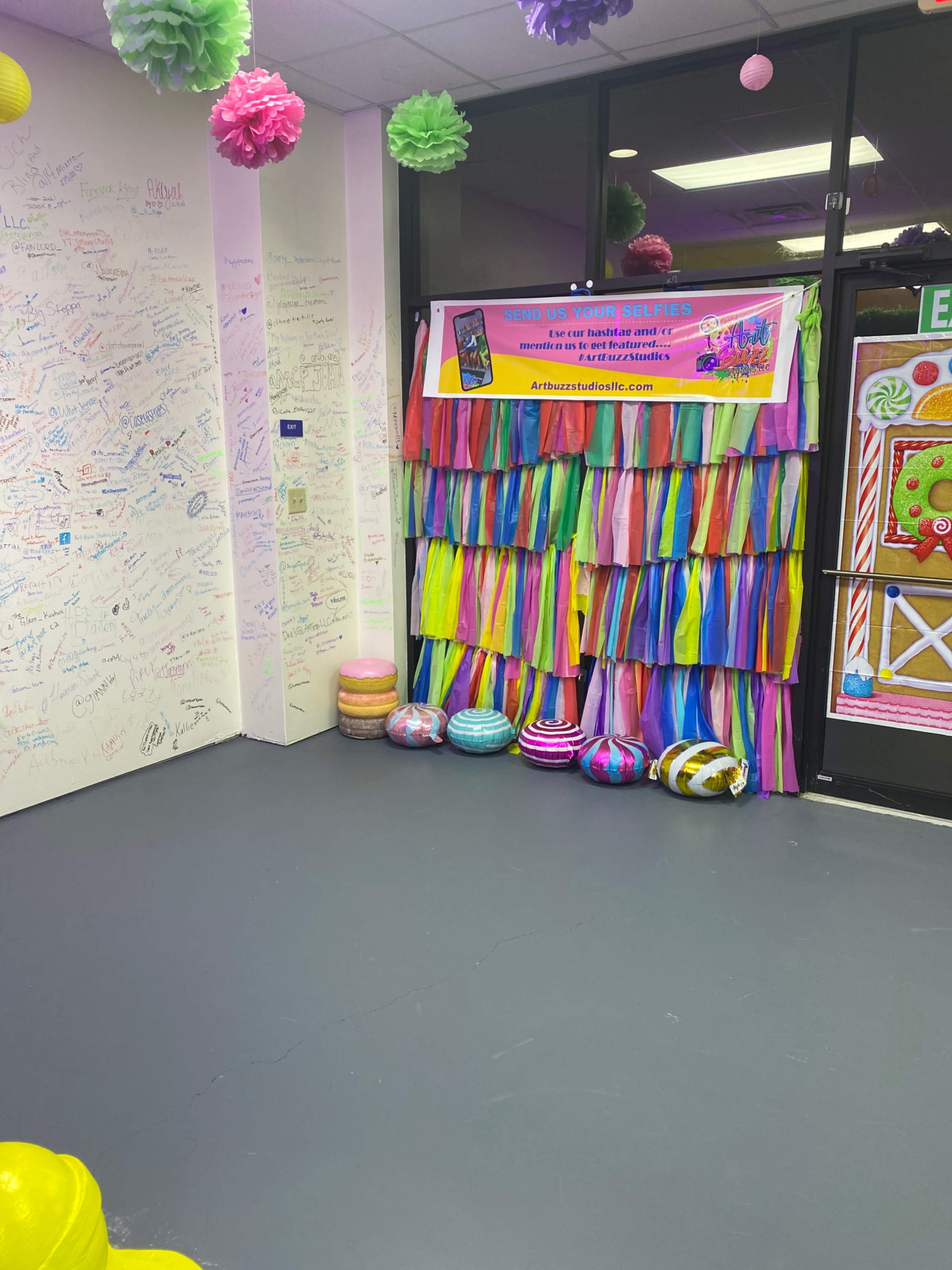 A brightly decorated corner with a colorful fringed backdrop, a promotional banner, and painted ornaments on the floor against a wall covered in signatures and drawings.