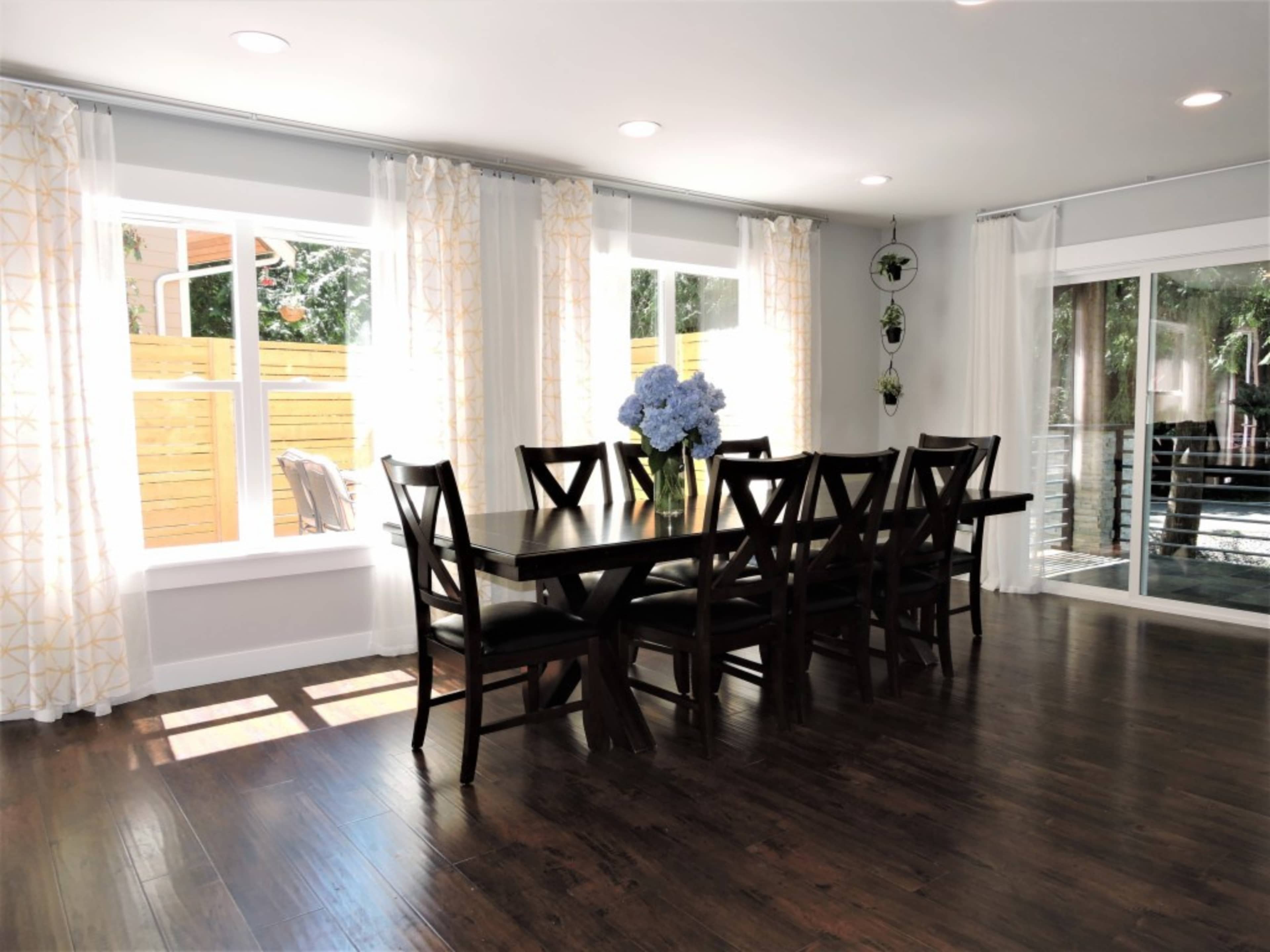 A spacious dining area features a large table surrounded by wooden chairs, with large windows allowing natural light and a view of the outdoor space.