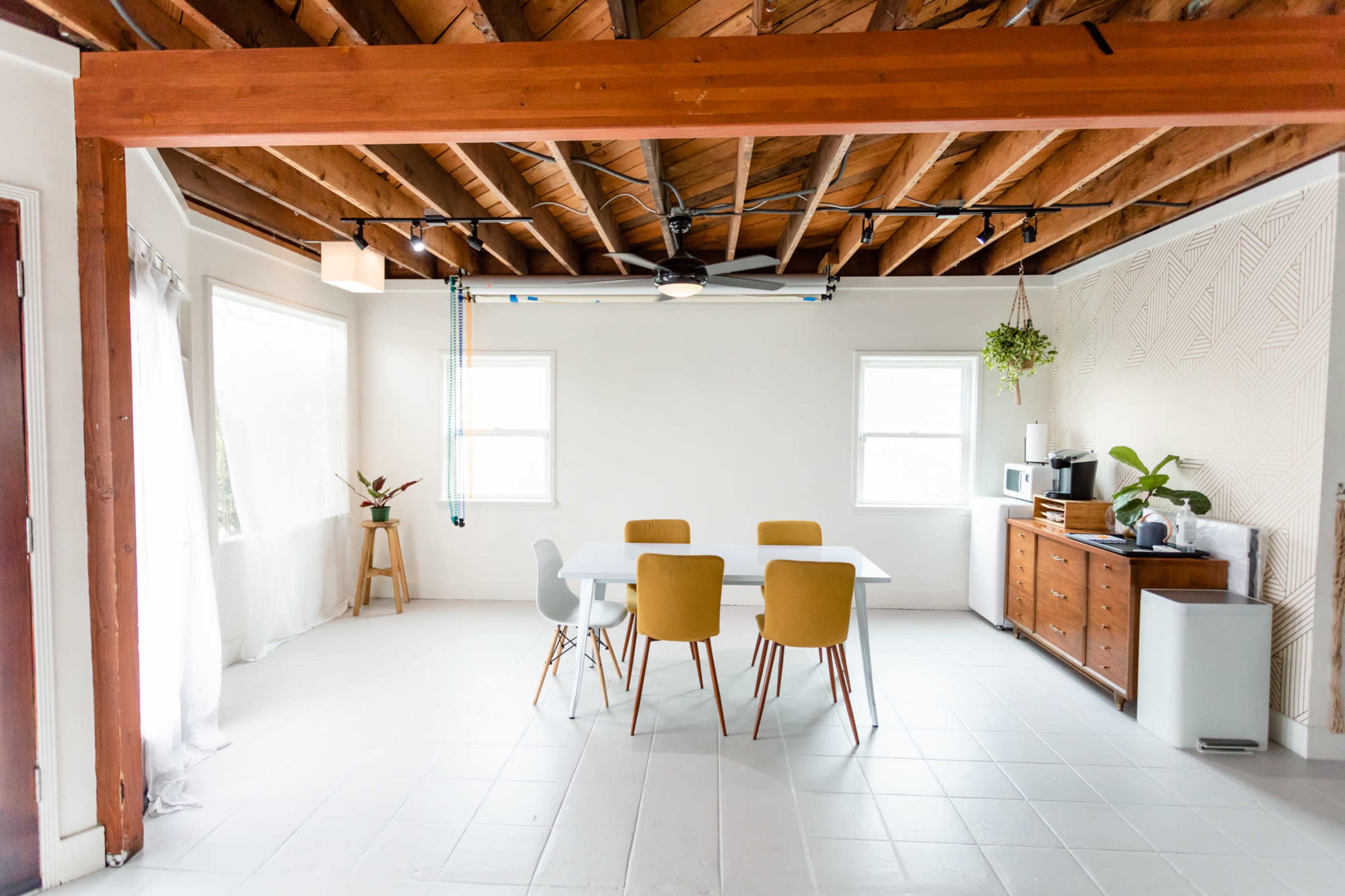 A minimalist dining area with a white table and four chairs surrounded by exposed wooden beams and light-colored walls.