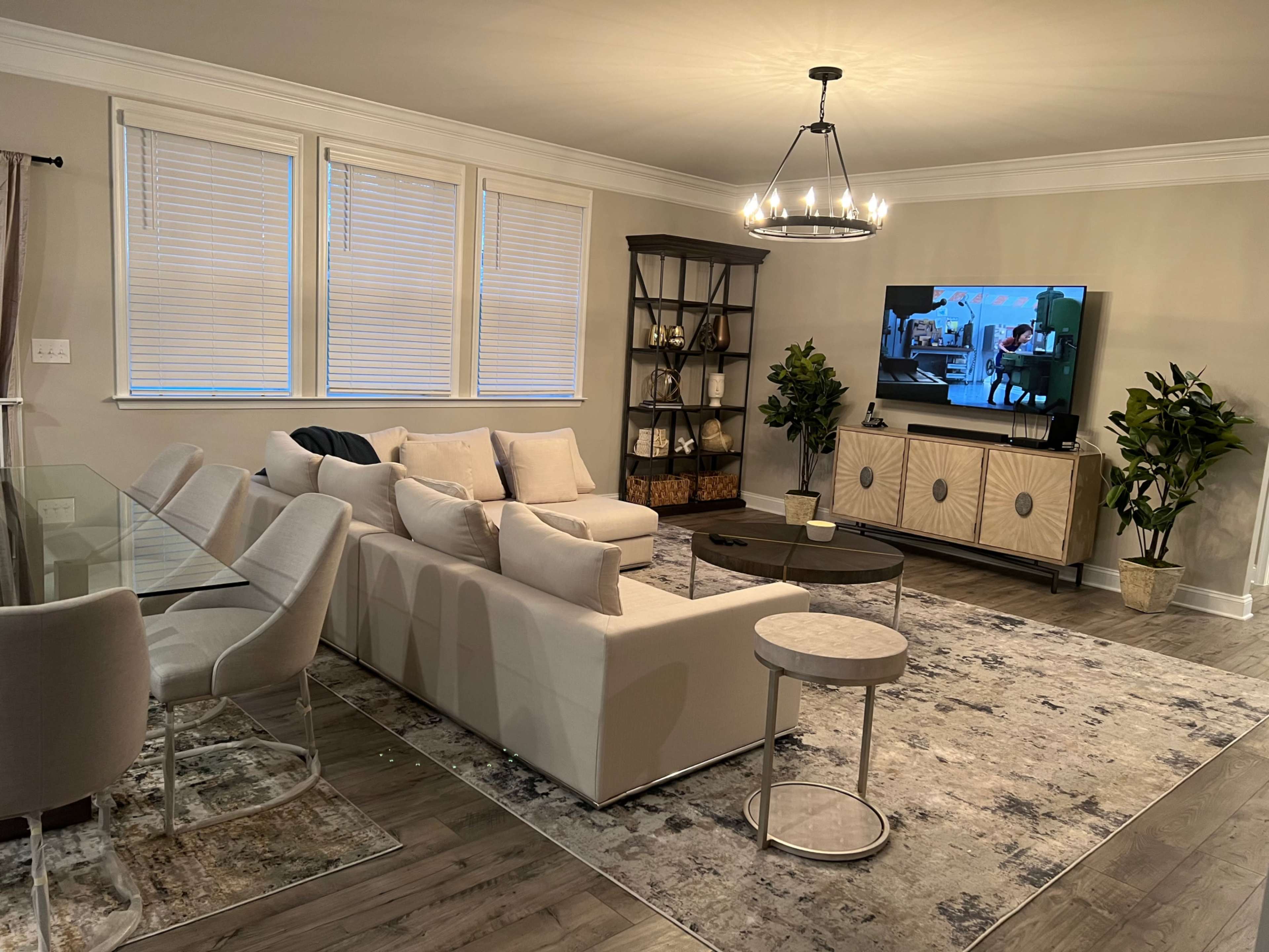 A modern living room featuring a sectional sofa, a glass dining table with chairs, indoor plants, and a television on a media console against a light-colored wall.