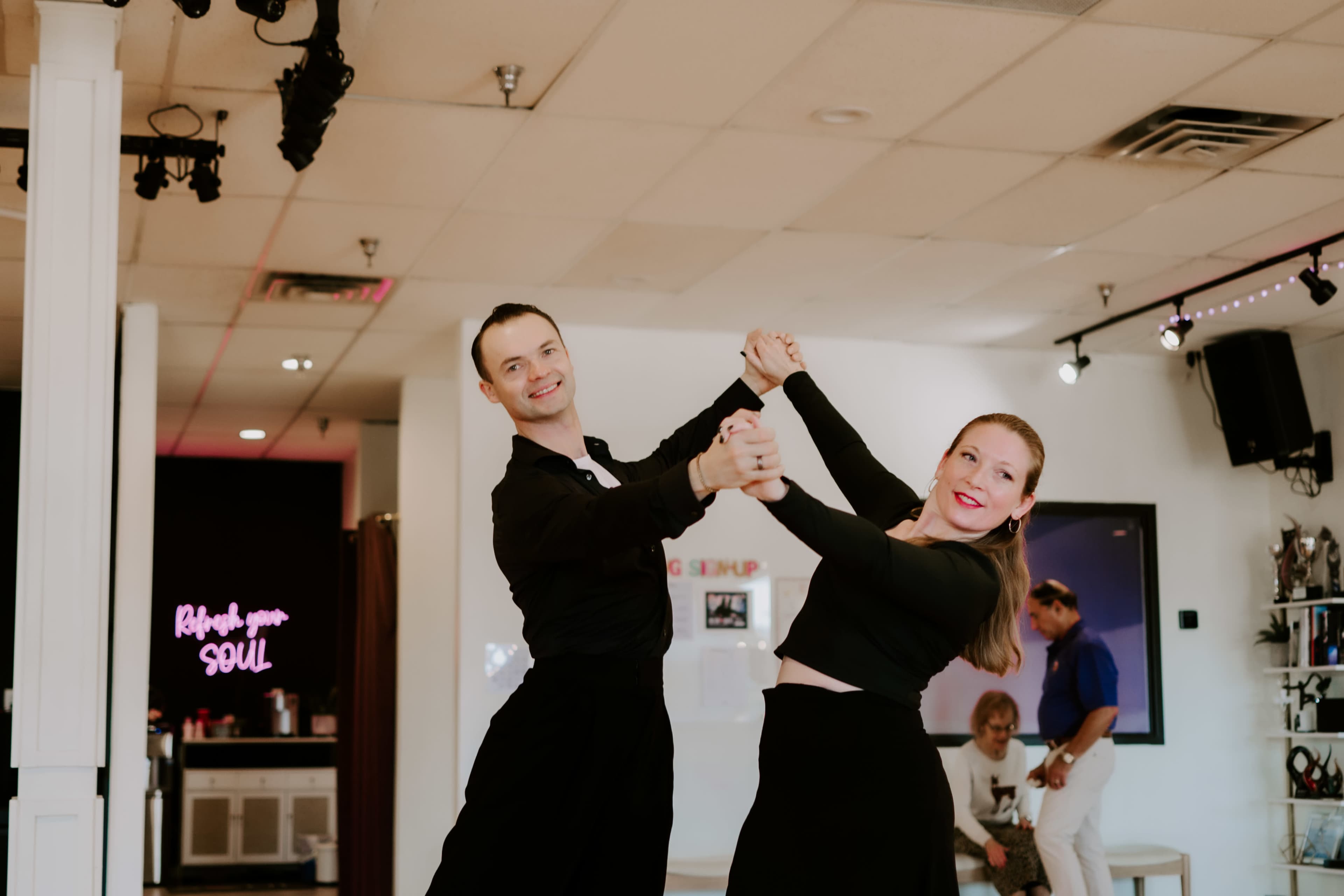 A couple dances together in a brightly lit studio with a neon sign in the background.