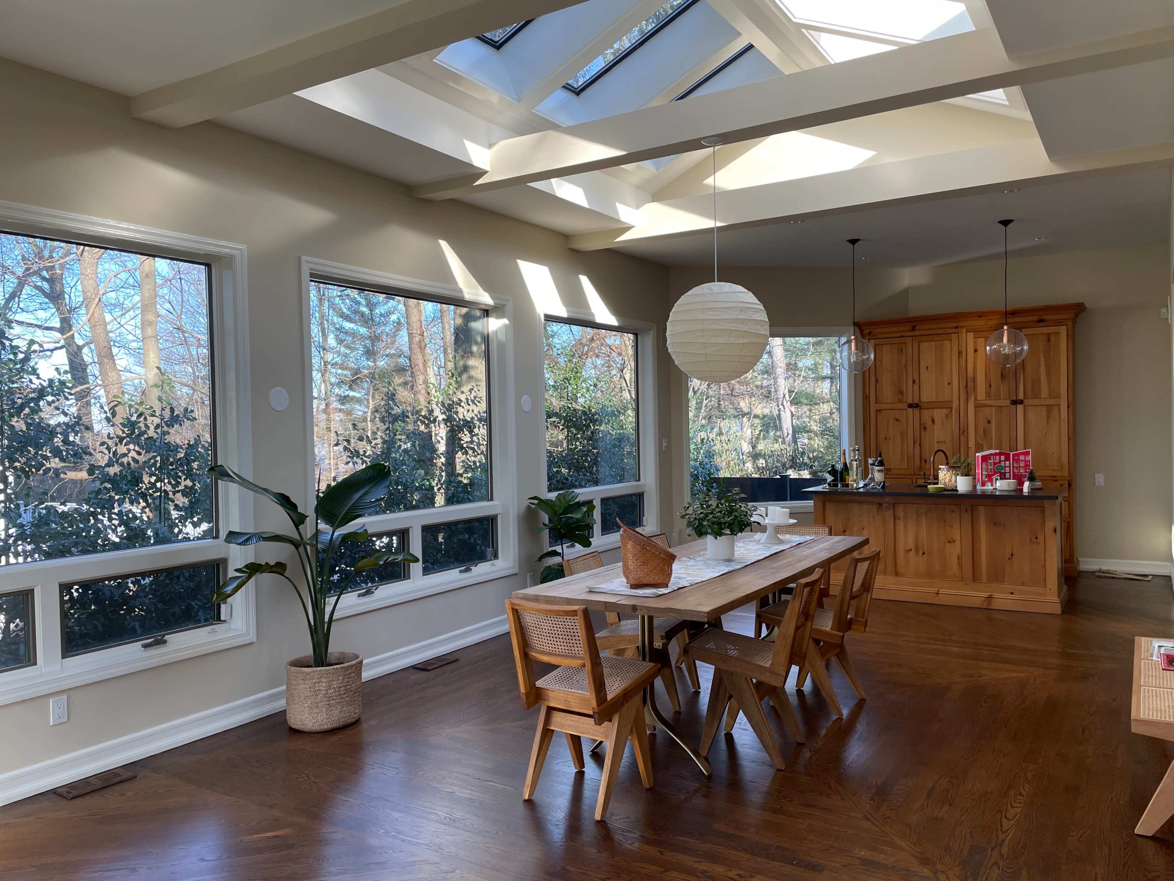 A spacious dining area features a wooden table surrounded by chairs, with large windows offering views of greenery and a wooden cabinet in the background.