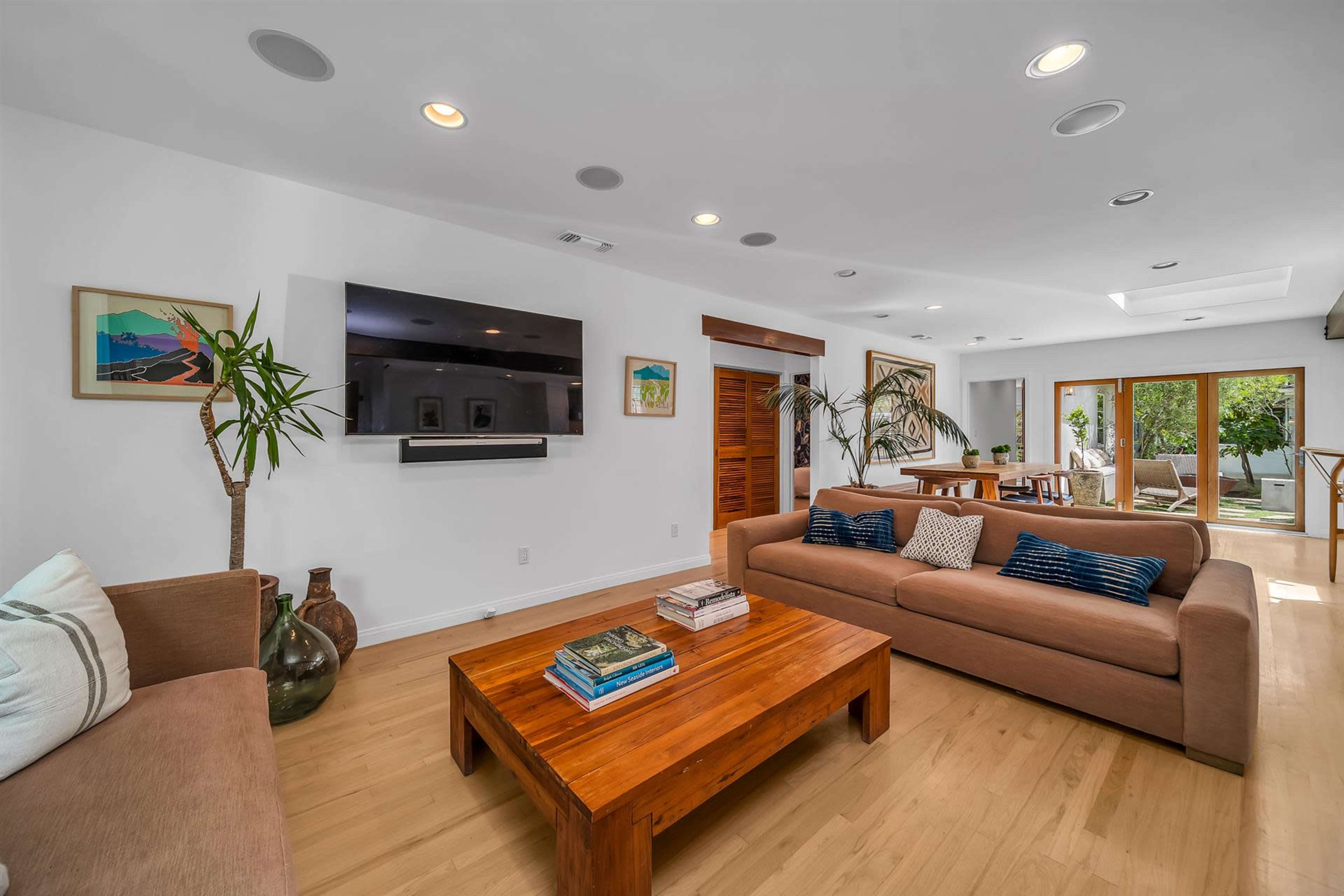The living room features a pair of sofas facing a wooden coffee table and a wall-mounted television, with large windows that open to a patio area.