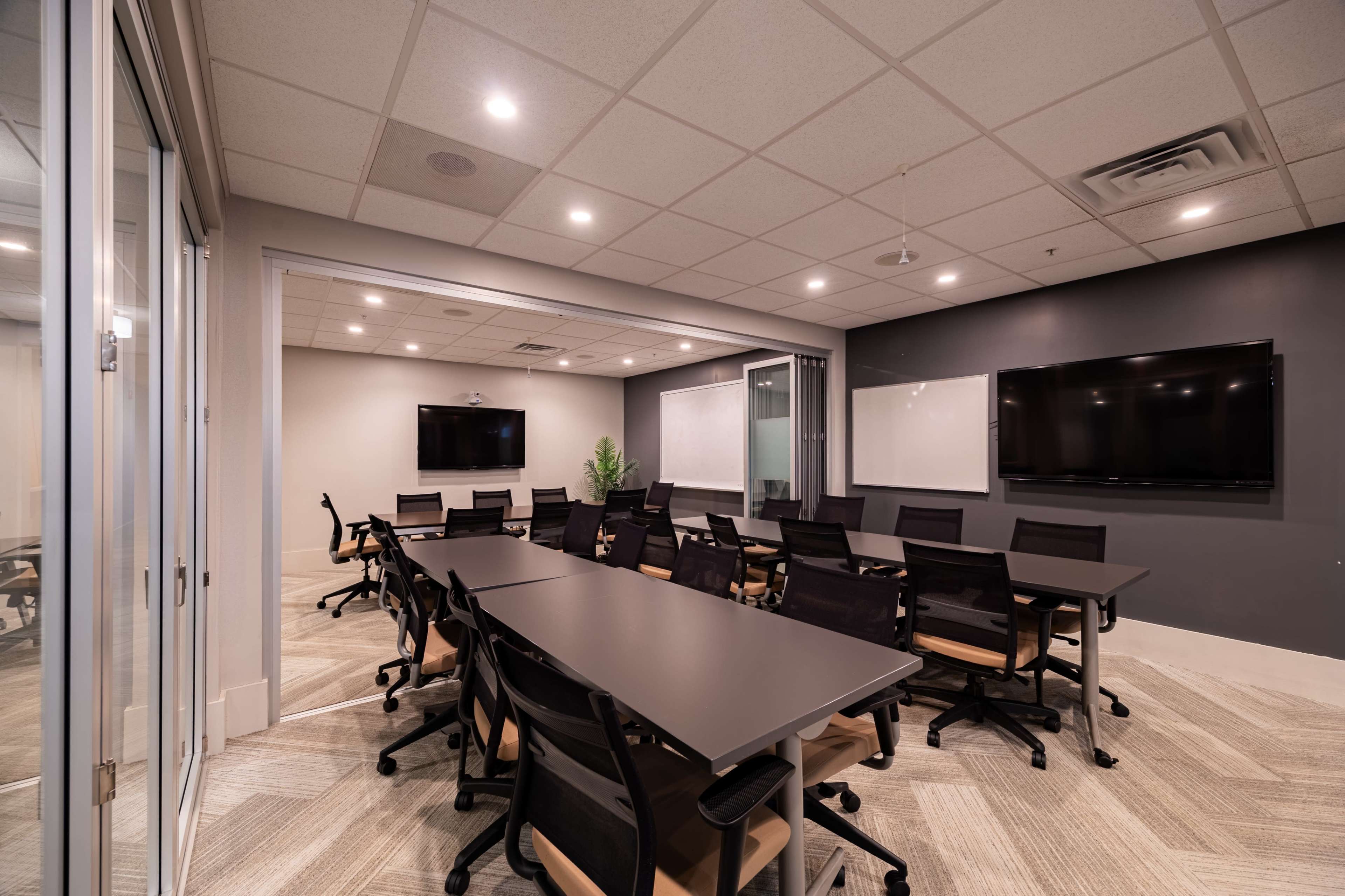 The image shows a modern conference room with a large table, multiple black chairs, two wall-mounted screens, and a plant placed in a corner.