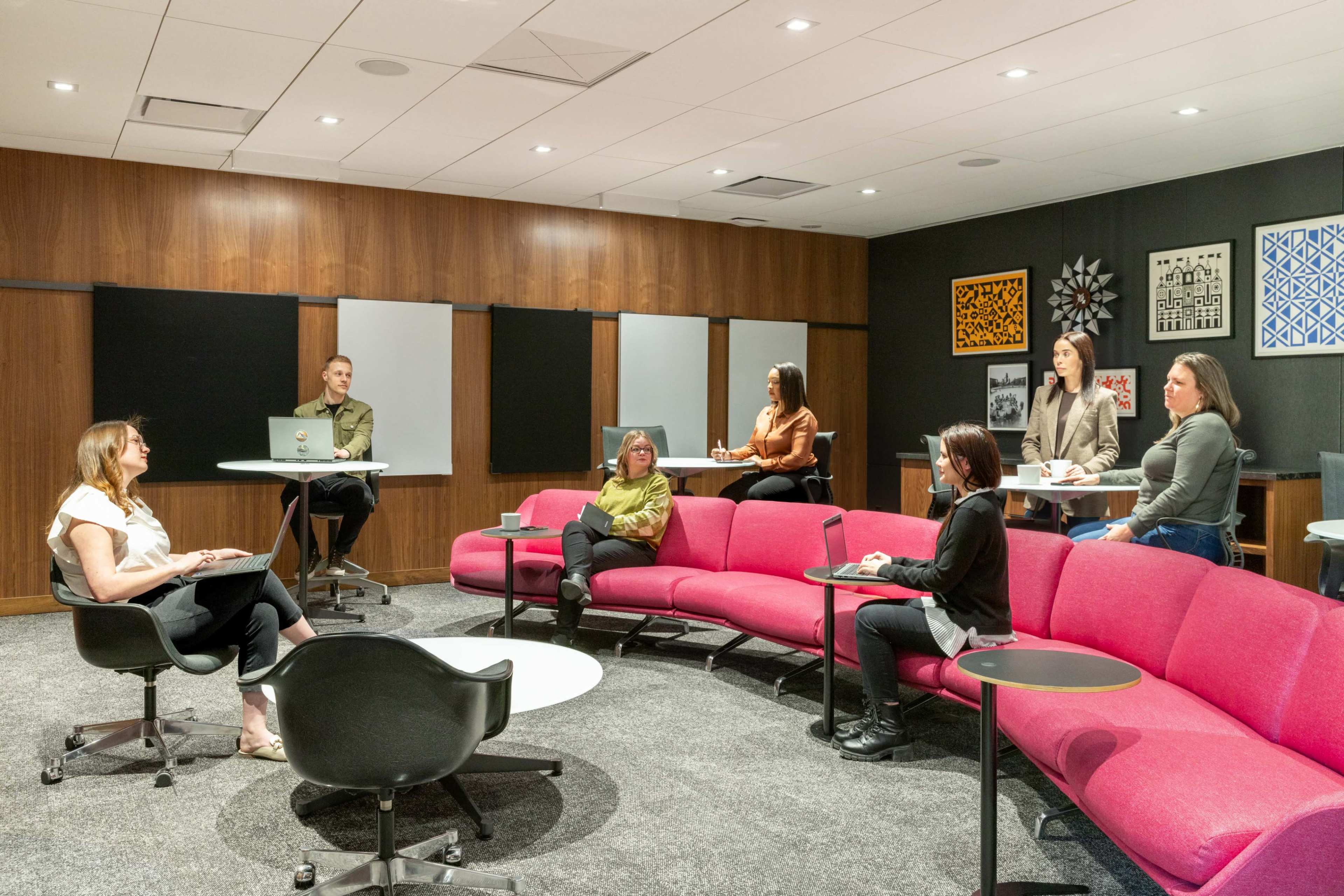 A group of six people is gathered in a modern meeting space featuring curved pink seating and a large screen, where one person is seated with a laptop while others stand or sit nearby.