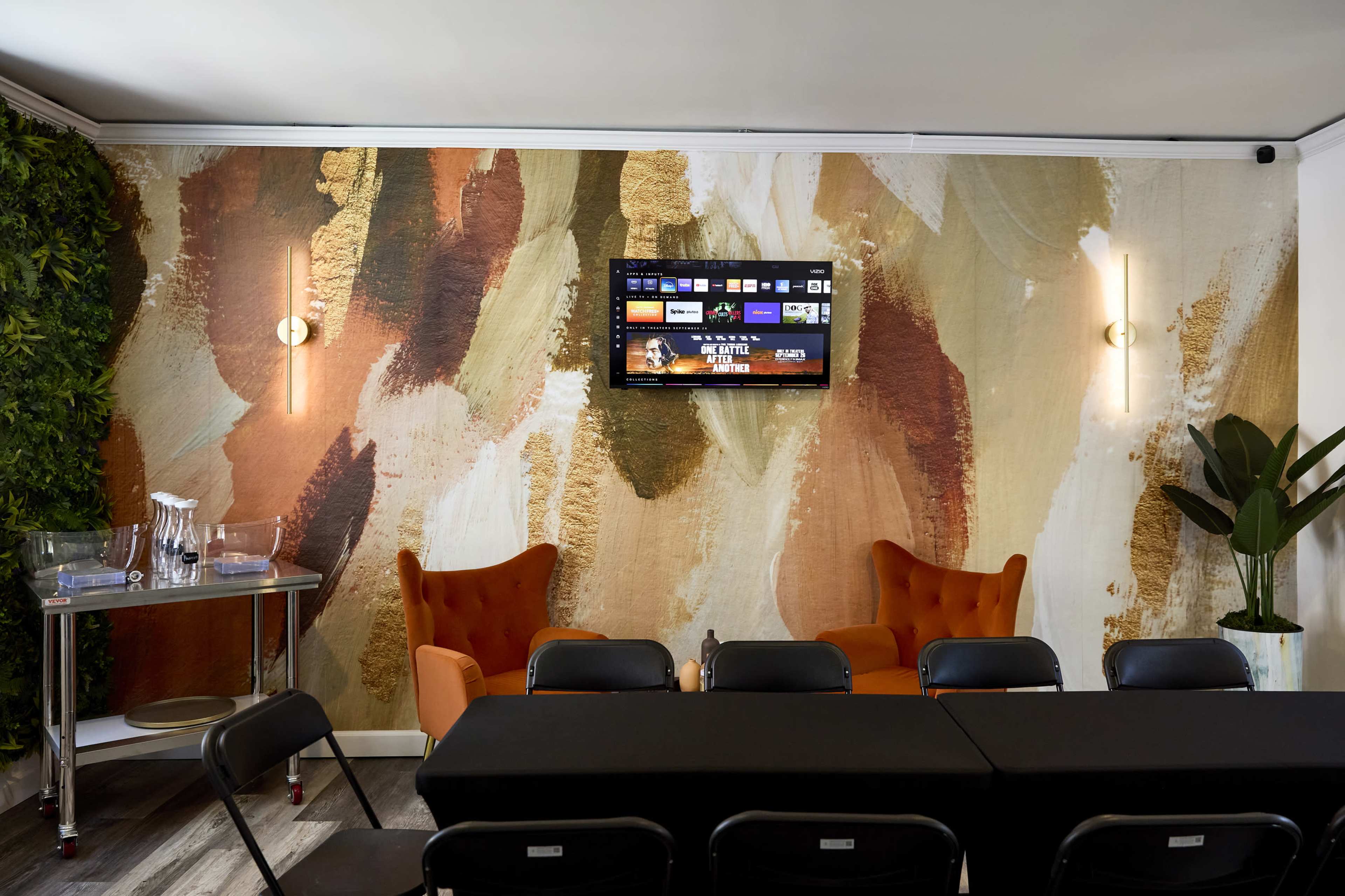 The image shows a modern interior with an abstract wall design, orange armchairs, a table with black chairs, and a wall-mounted television.