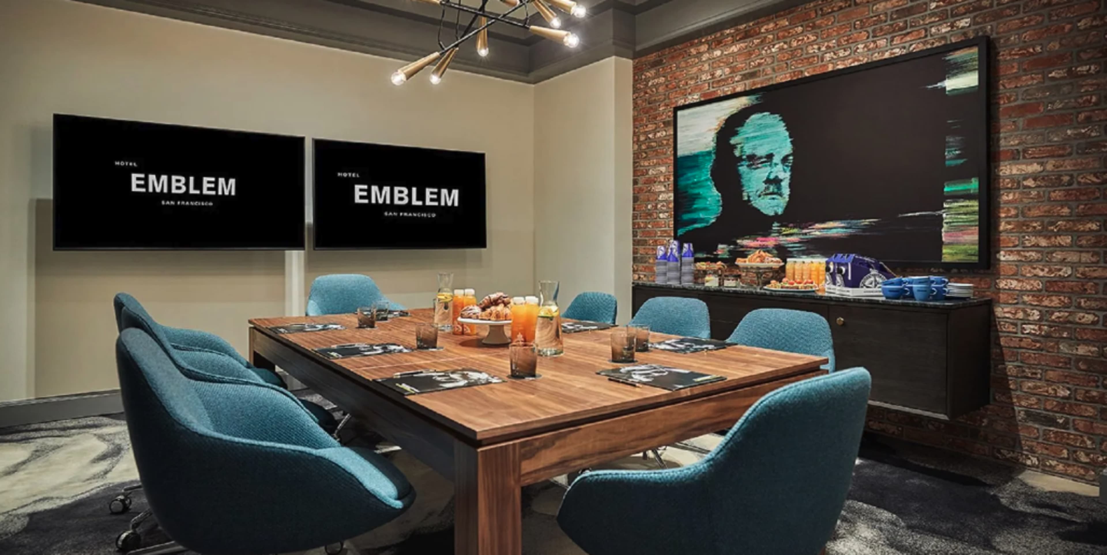 A conference room features a wooden table surrounded by teal chairs, with two screens displaying the hotel name and a large painting on the wall.