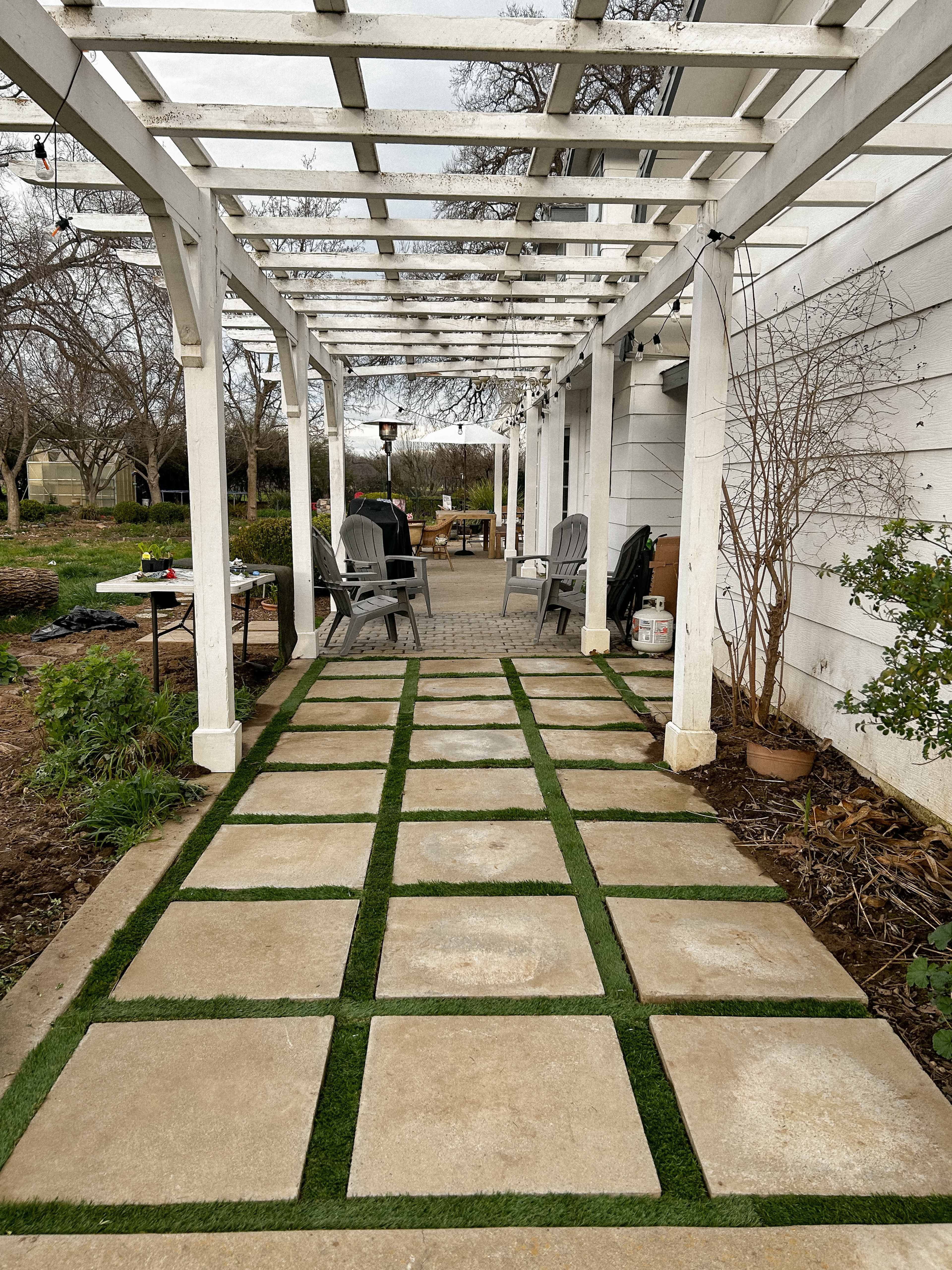 The image shows a pathway made of large stone slabs interspersed with patches of grass, leading toward a patio with outdoor seating under a wooden trellis.