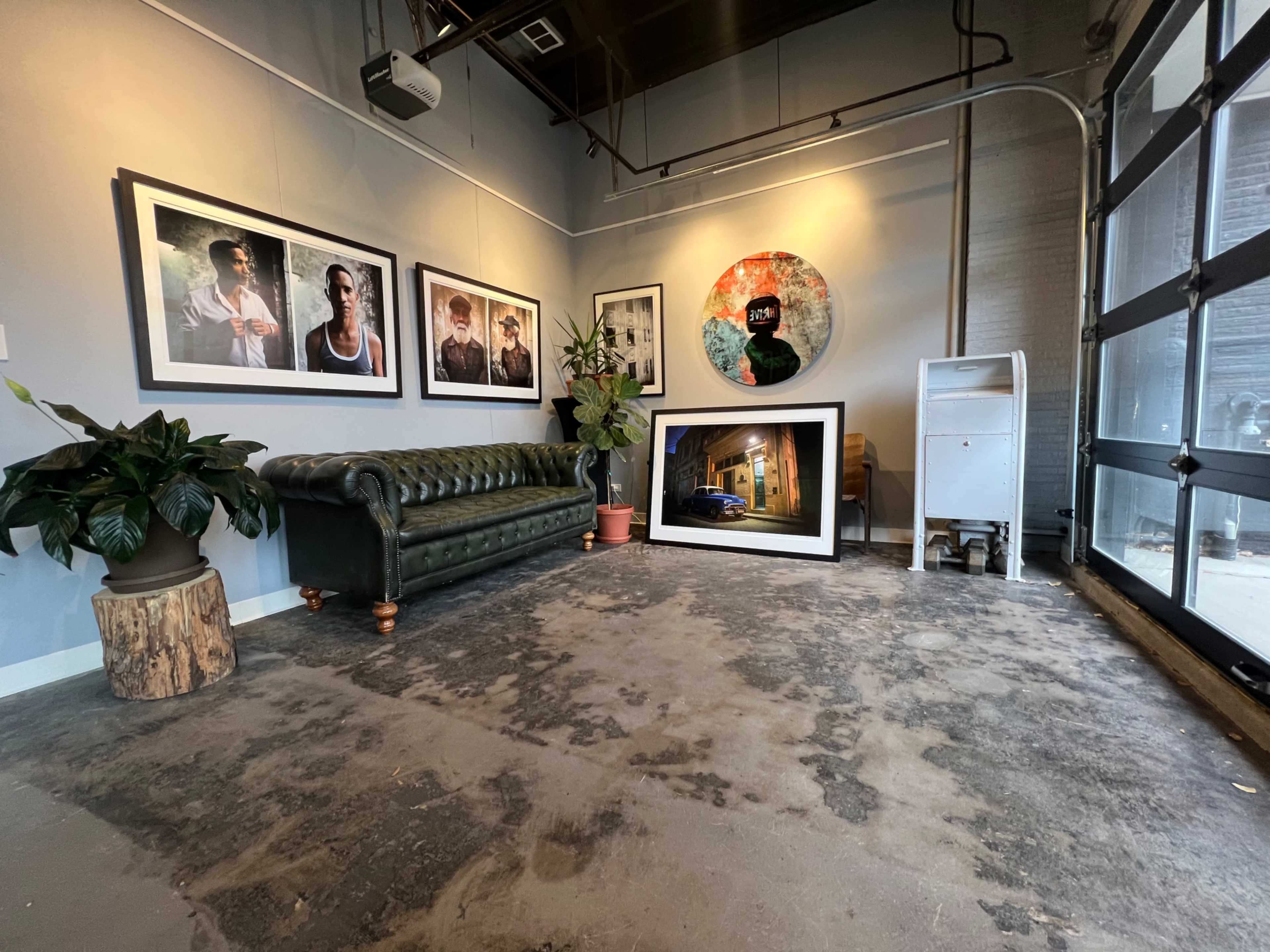 The image shows a spacious art gallery with a green leather sofa, framed photographs on the walls, a large circular artwork, and a leaning framed picture on the floor.