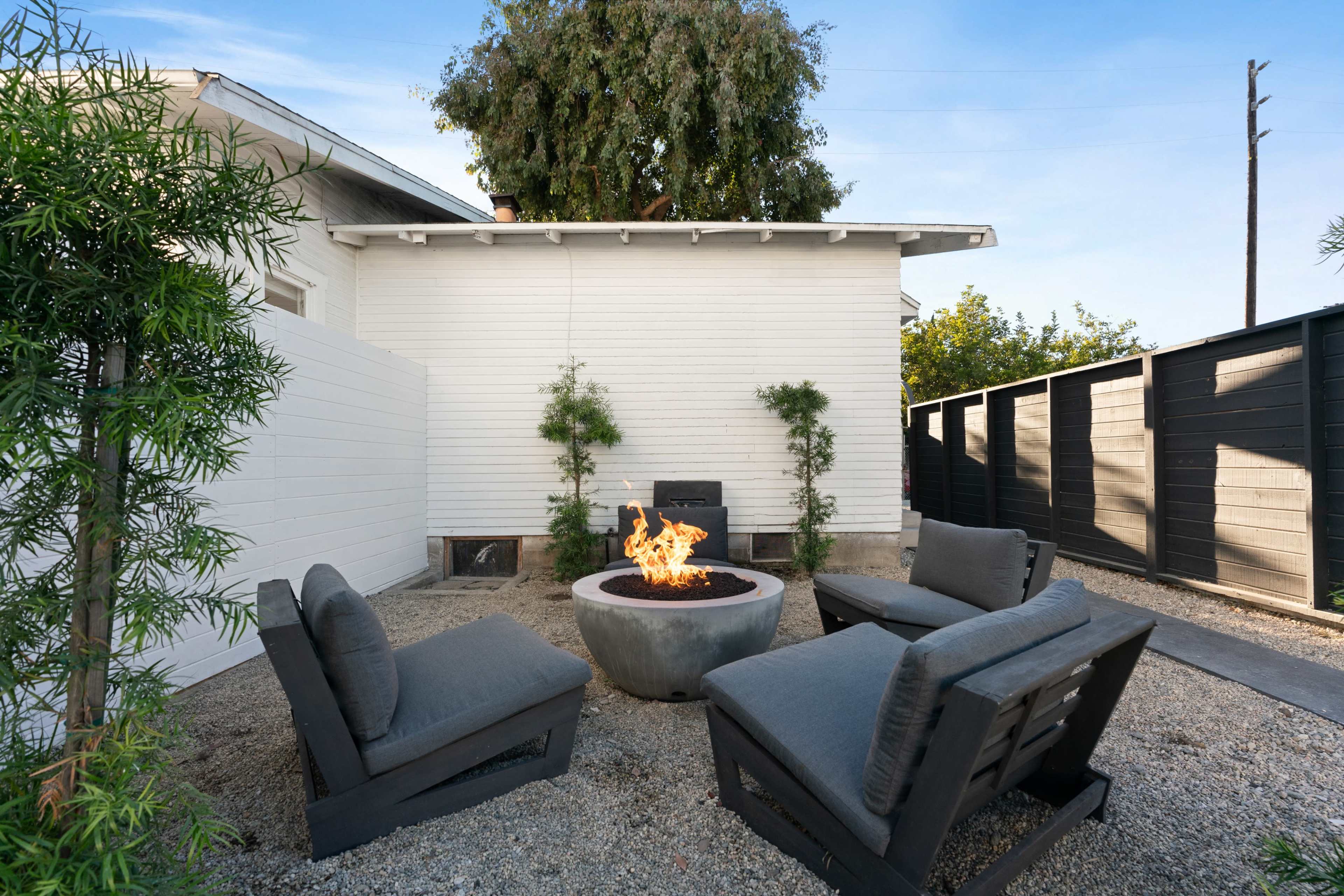 The image shows a cozy outdoor seating area with four chairs arranged around a fire pit, surrounded by gravel and green plants.