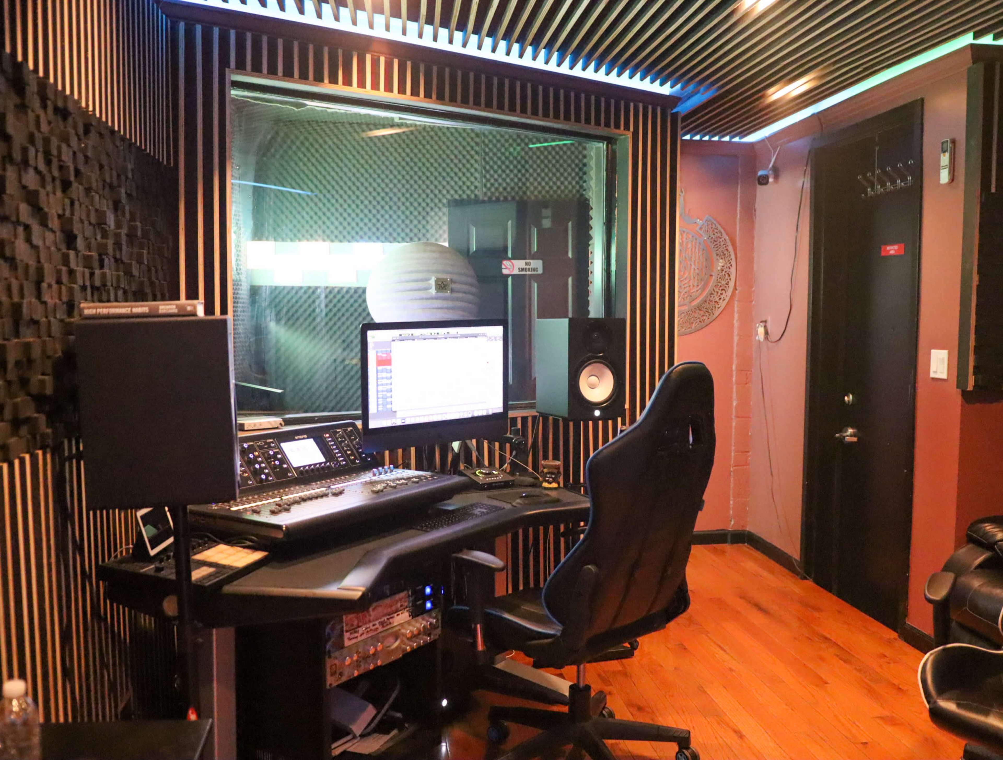 The image shows a recording studio with a computer setup, sound equipment, and a black office chair in a room with soundproof walls.