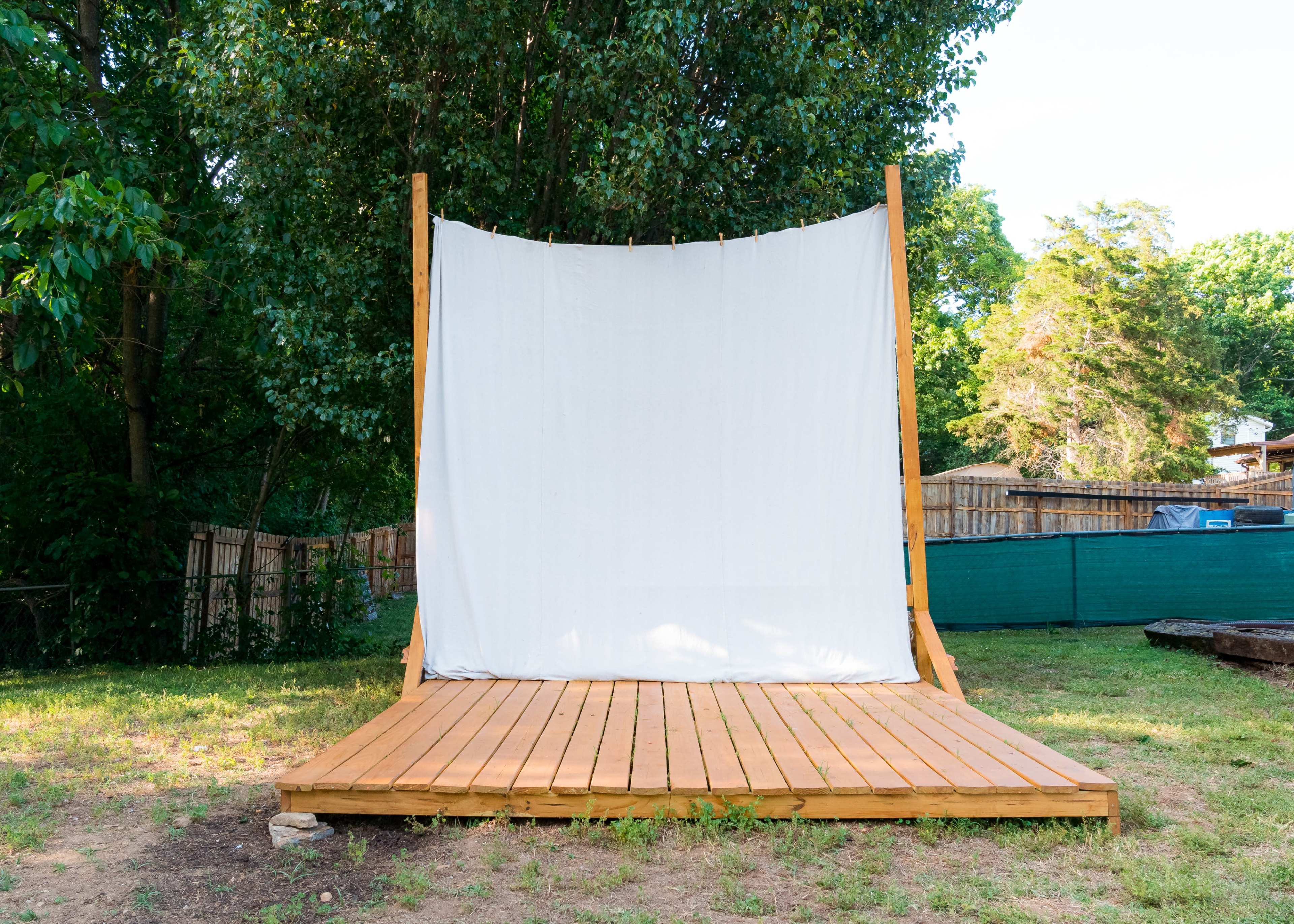 The image shows a wooden platform with a large white backdrop set up in a green outdoor area surrounded by trees.
