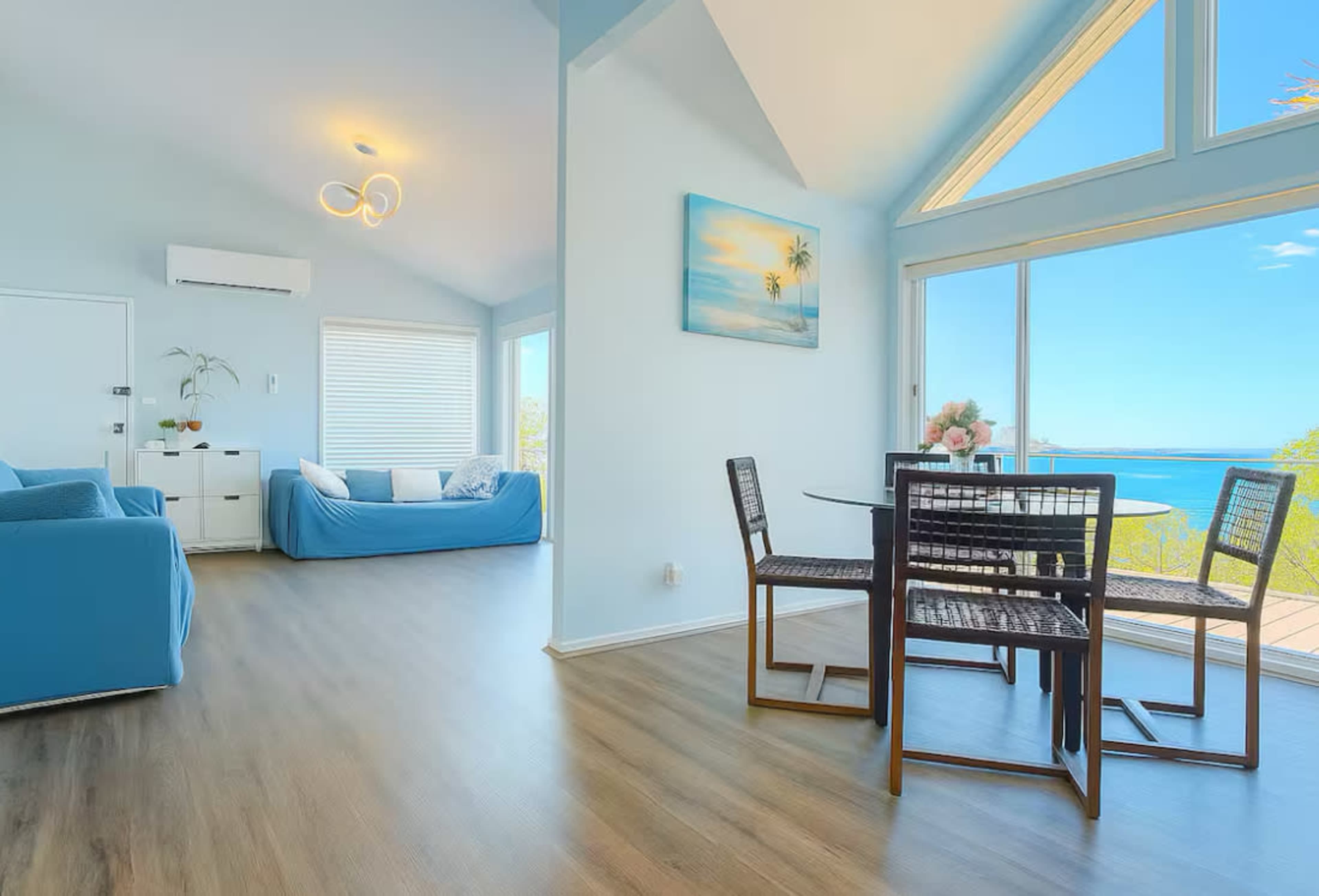 The image shows a bright, open living area featuring a blue sofa, a dining table with chairs, and large windows offering a view of the ocean.