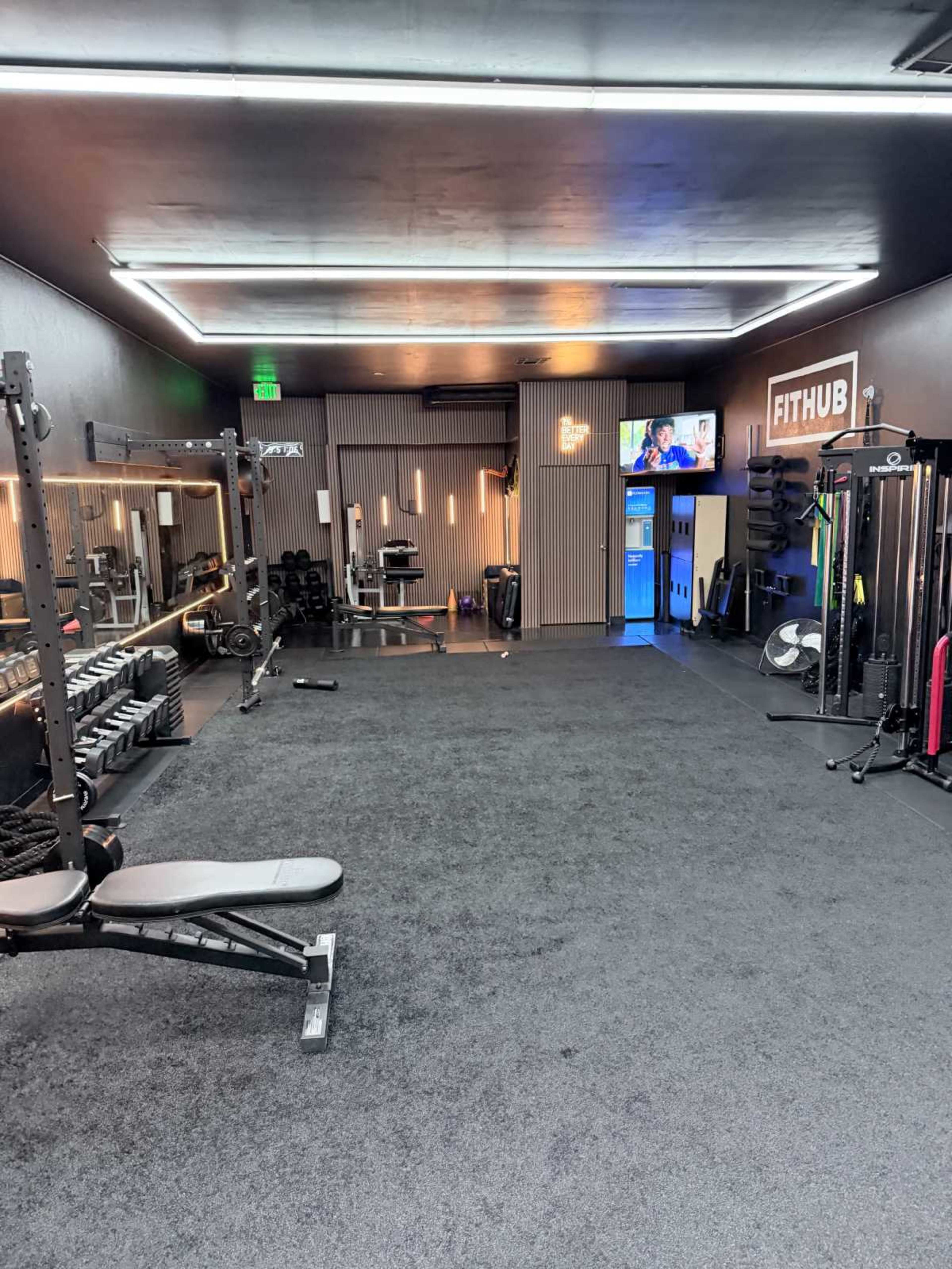 The image shows a modern gym interior with equipment like weights, benches, and cardio machines, featuring dim lighting and a television on one wall.