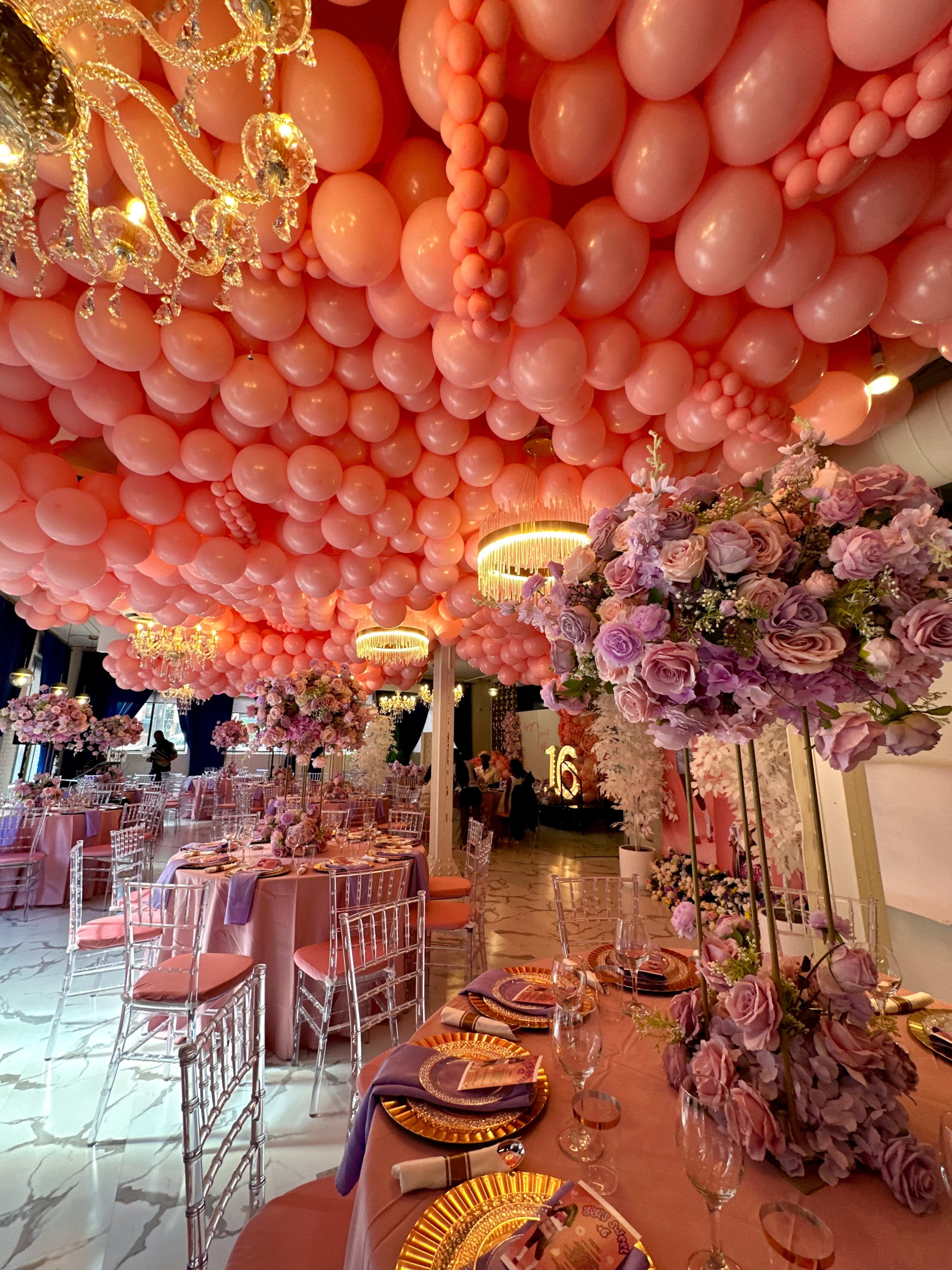 The scene features an elegantly decorated event space with a ceiling covered in pink balloons, tables adorned with floral centerpieces, and lavish place settings.