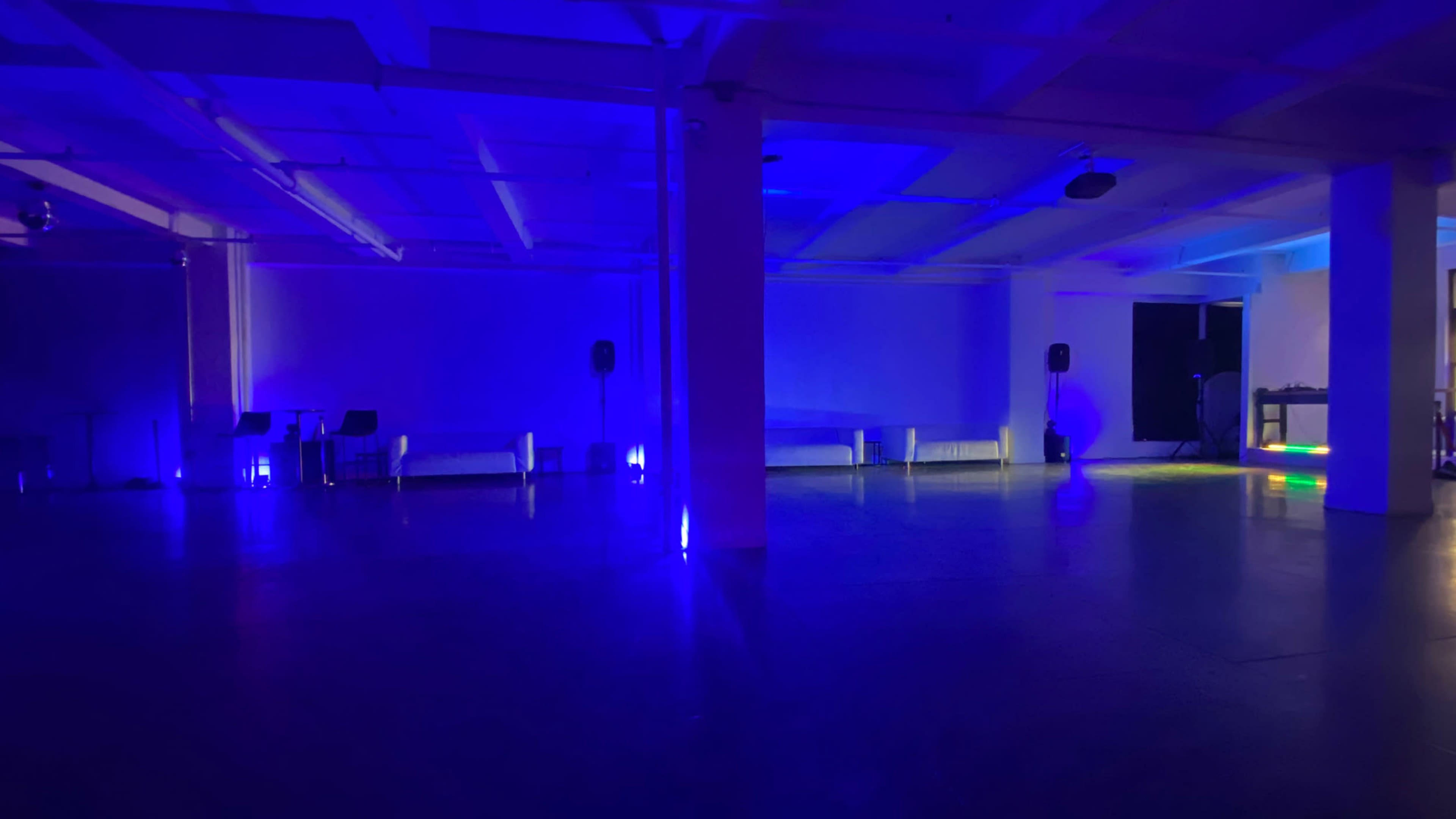 An empty room with blue lighting, featuring a polished floor and sparse seating arrangements along the walls.