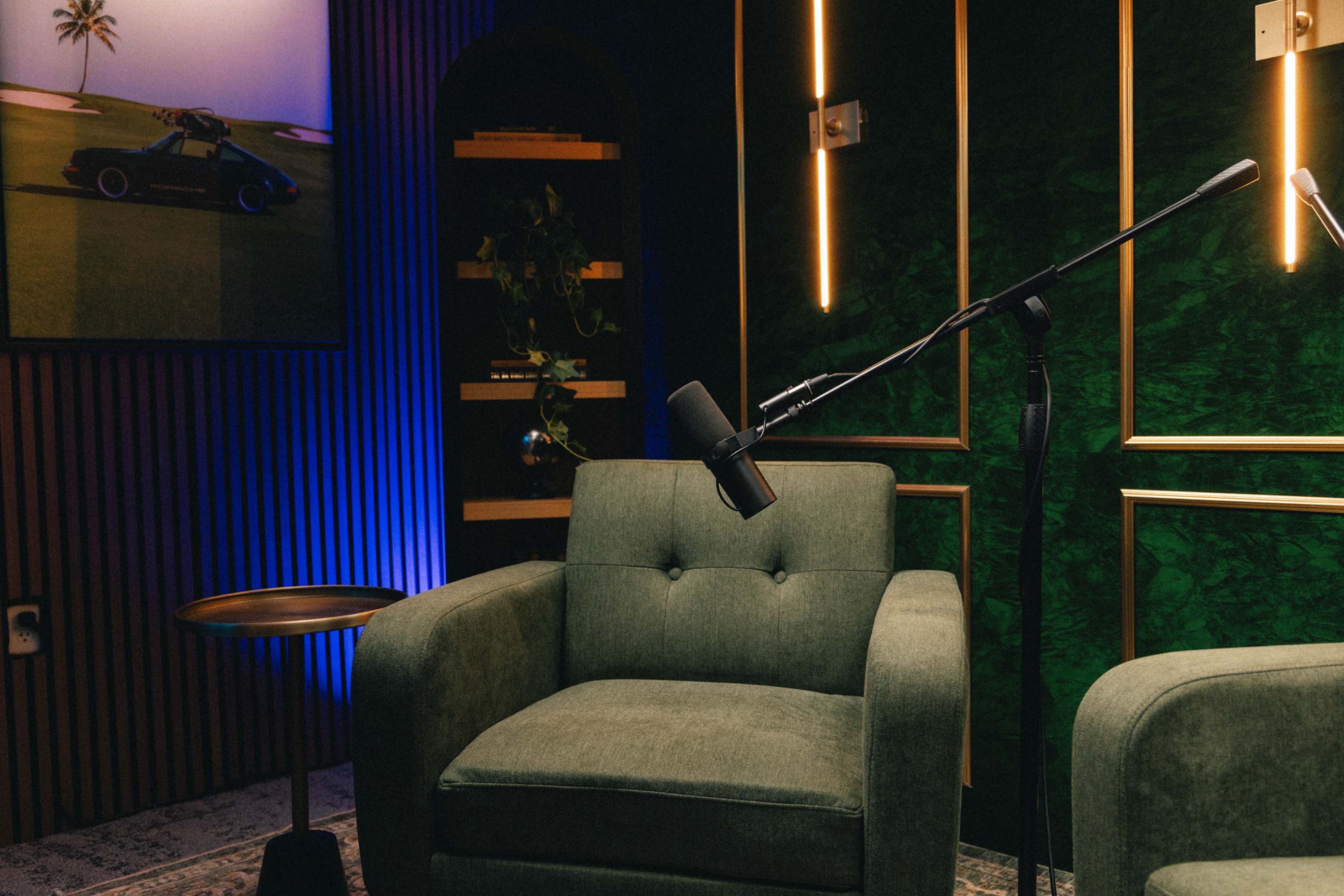 The image shows a cozy interior featuring a green armchair, an adjacent wooden side table, and a microphone set up for recording, with decorative lighting and artwork in the background.