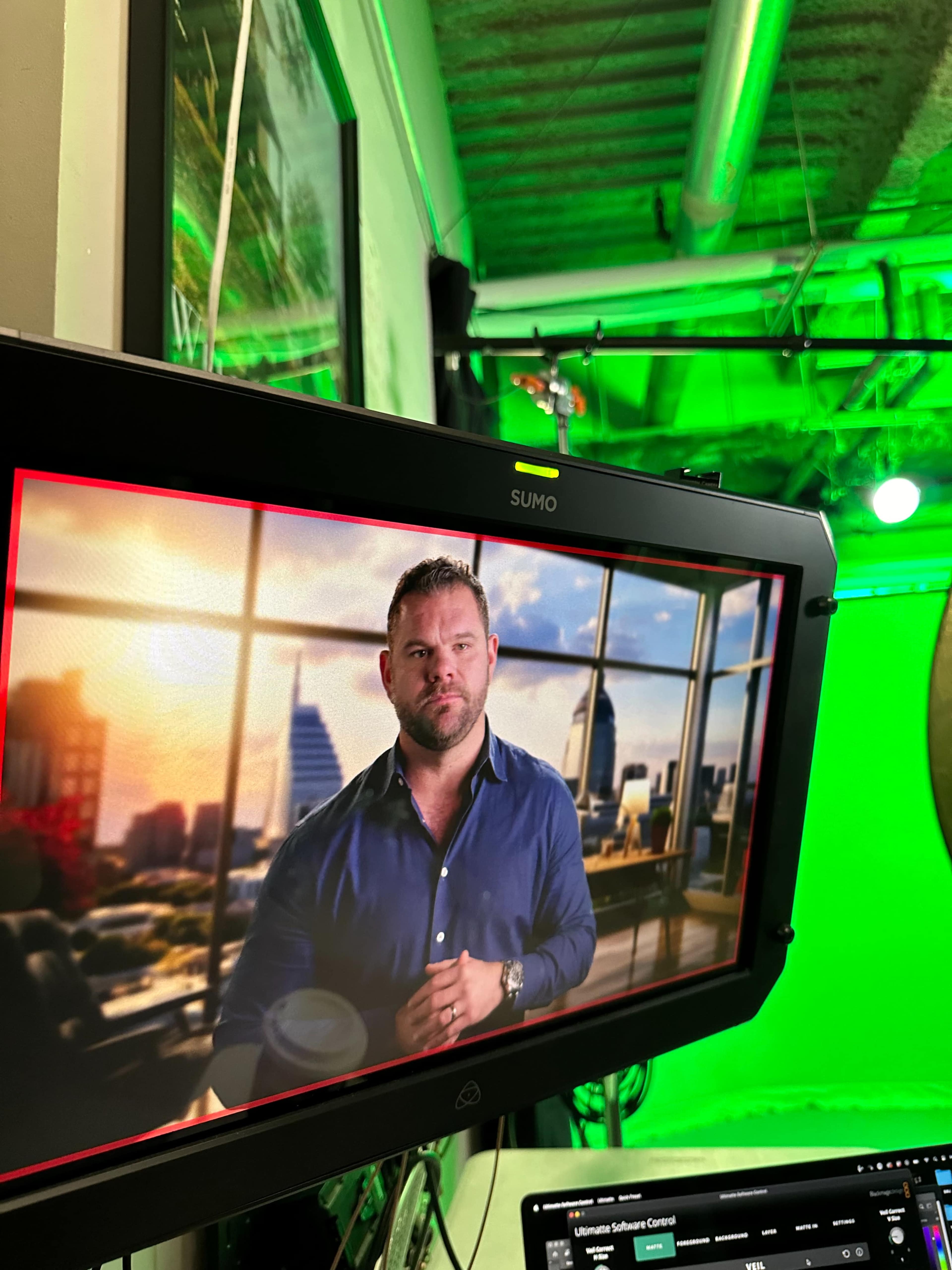 A man in a blue shirt stands in front of a city skyline displayed on a monitor, with a green screen in the background.