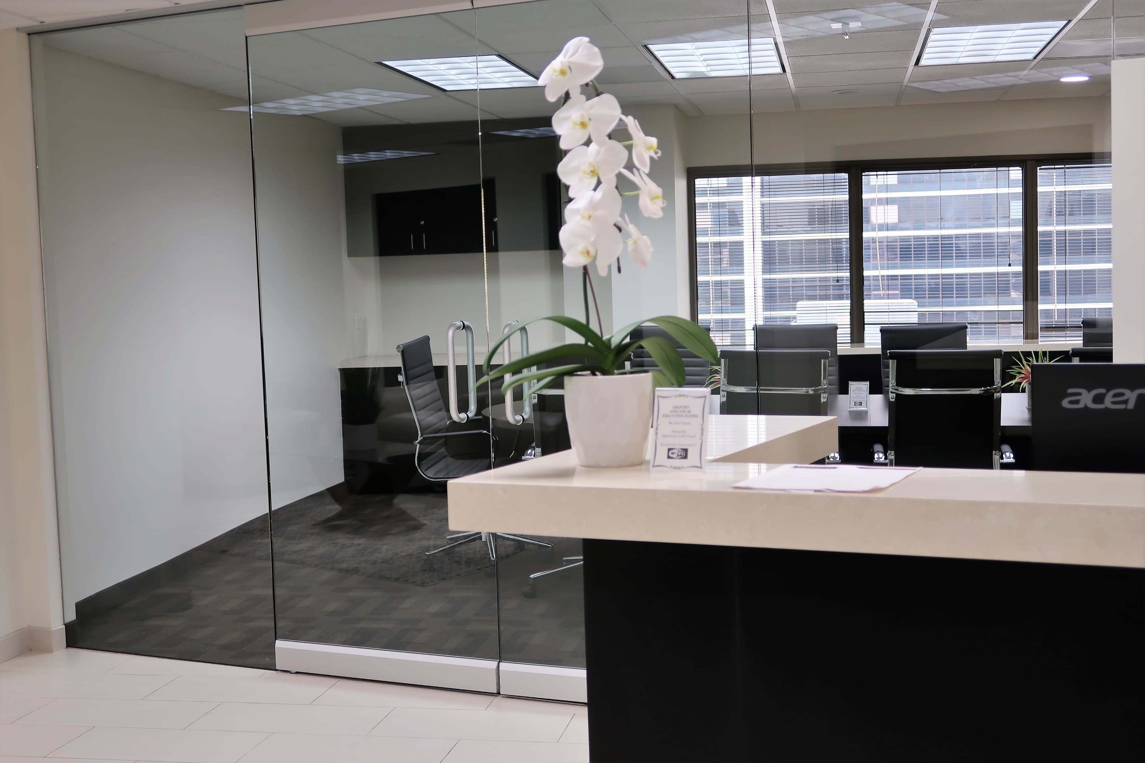 The image shows a modern office reception area with a large glass wall, a desk with a potted orchid, and a view into a conference room.