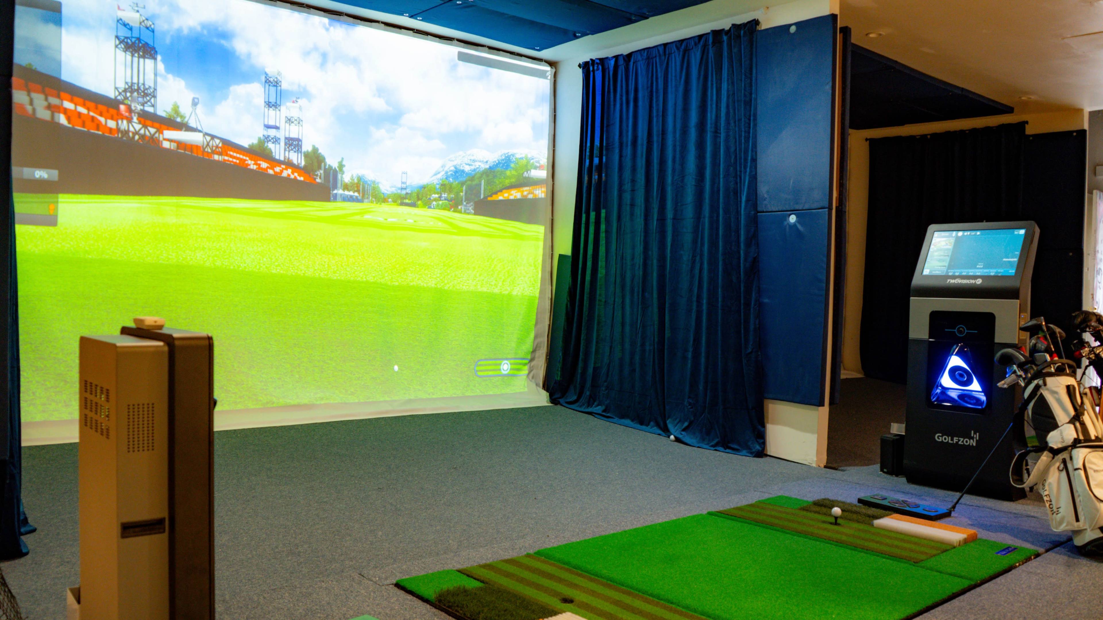An indoor golf simulator setup with a large projection screen displaying a virtual golf course, a putting mat, and golf clubs nearby.