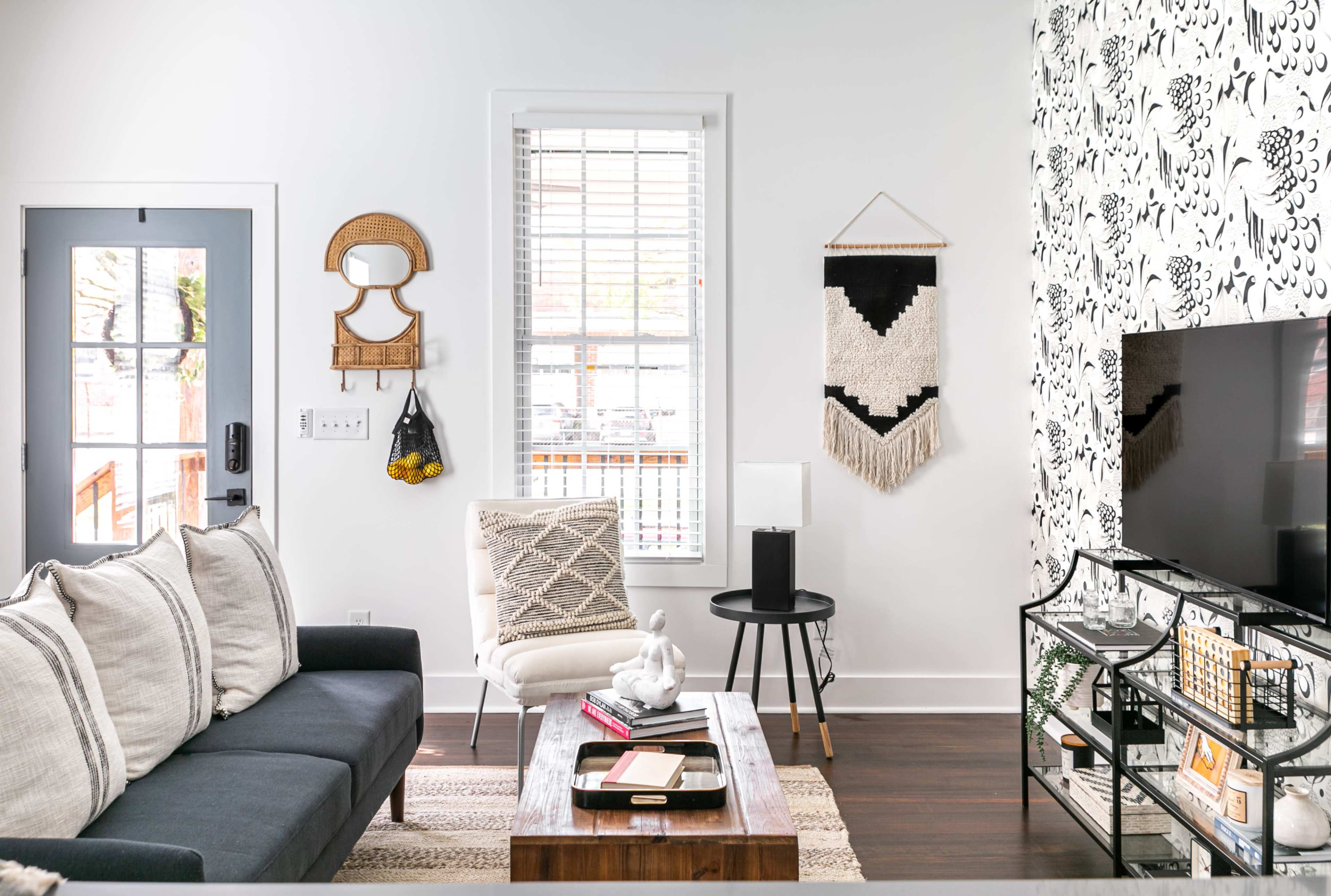 The living room features a dark sofa, a coffee table, a black and white patterned wall, and a television on a stand, with natural light coming through the windows.