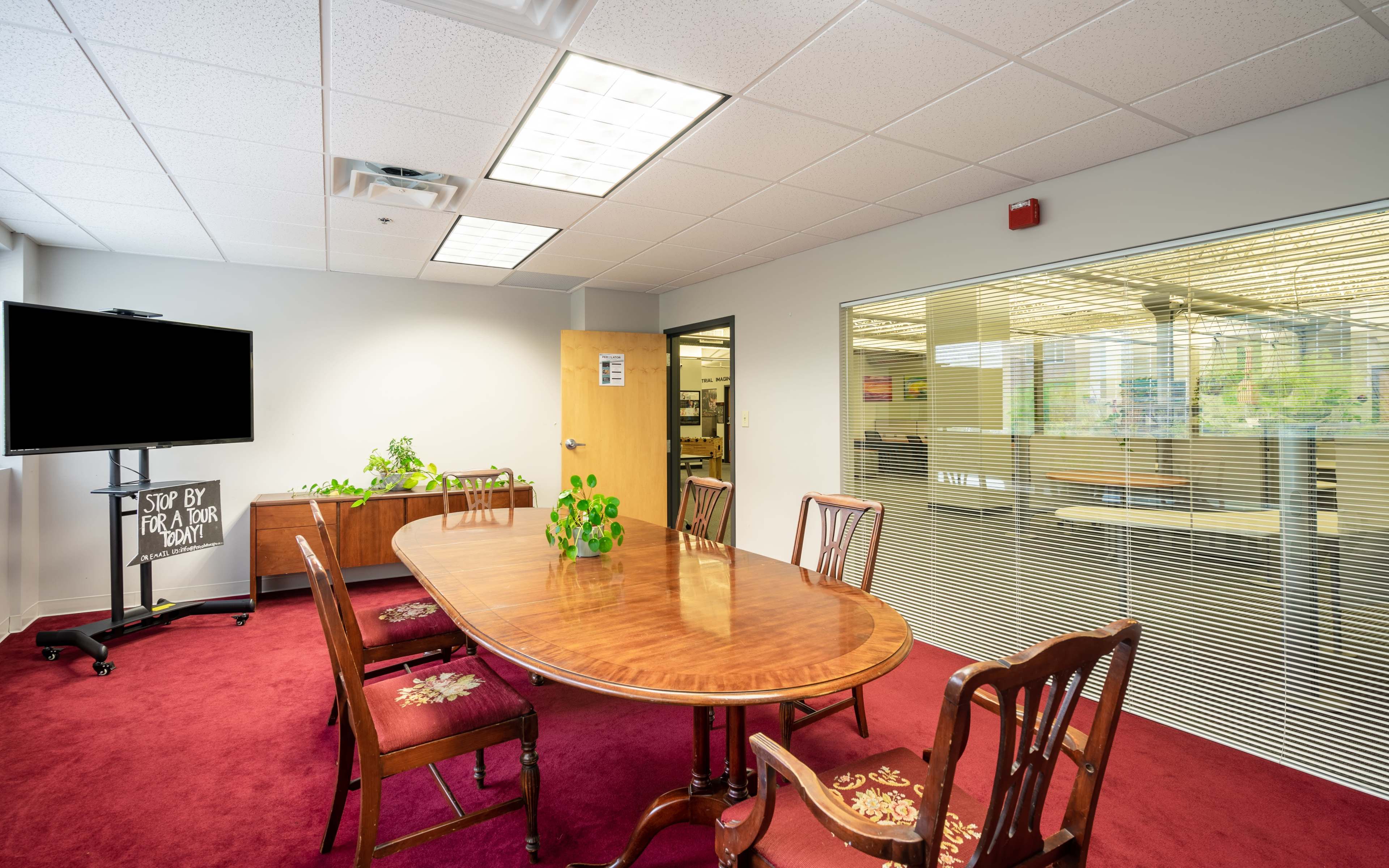 A conference room with a wooden table surrounded by chairs, a television on a stand, and large windows displaying an office space outside.