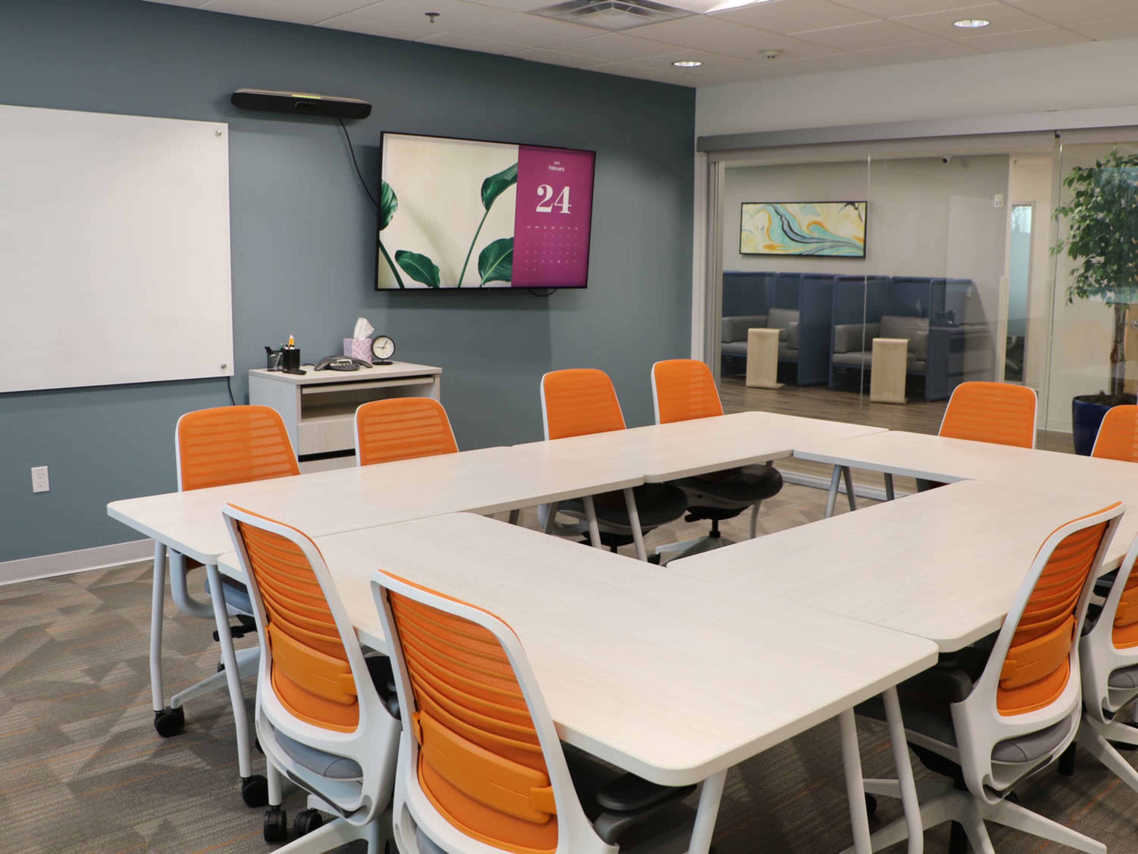 The image shows a modern conference room with a large white table arranged in a U-shape, surrounded by orange and white chairs, and equipped with a wall-mounted display and a whiteboard.