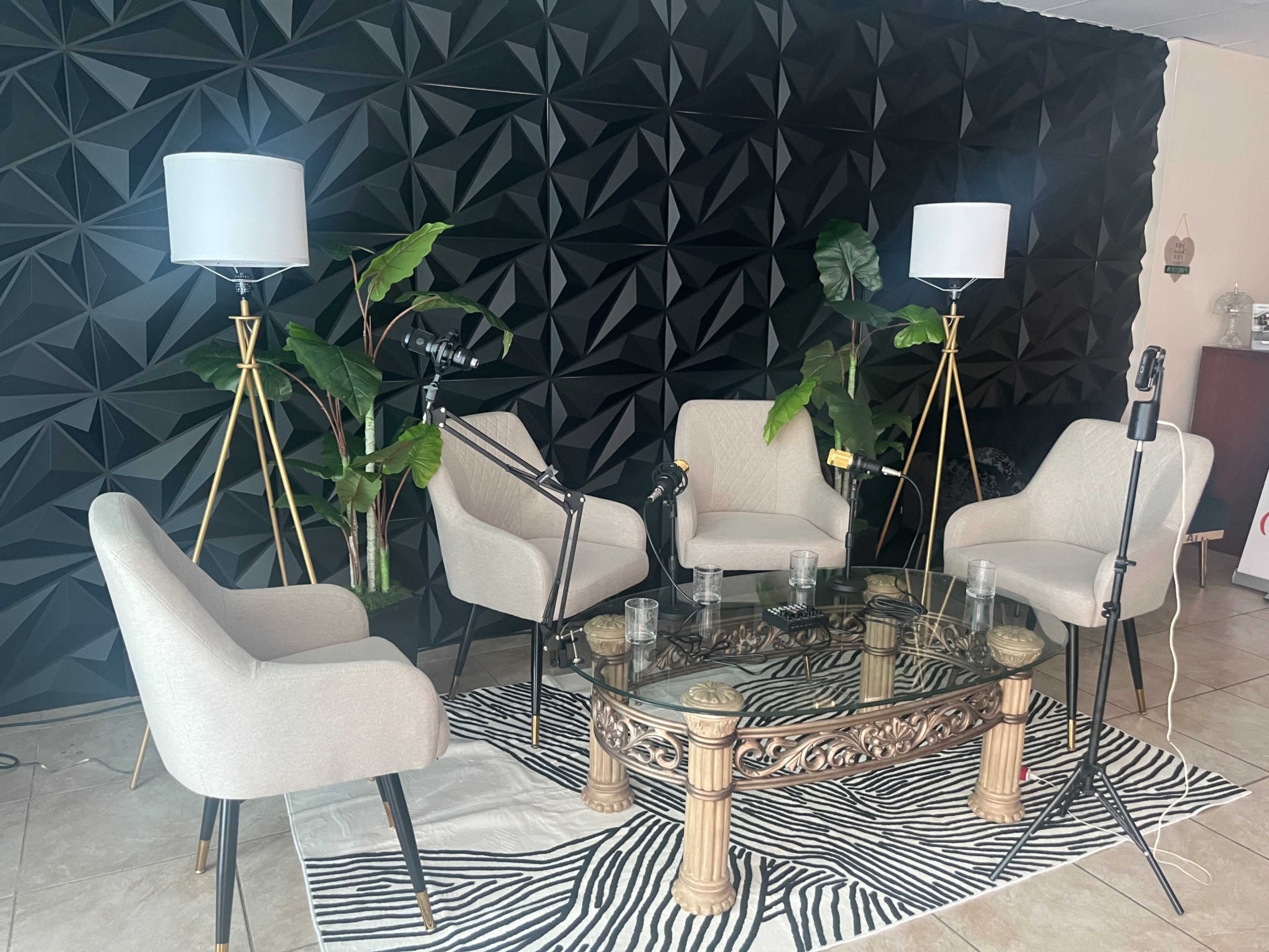 The image shows a seating area with four beige chairs arranged around a glass coffee table, set against a black 3D-patterned wall, with two floor lamps and a plant.