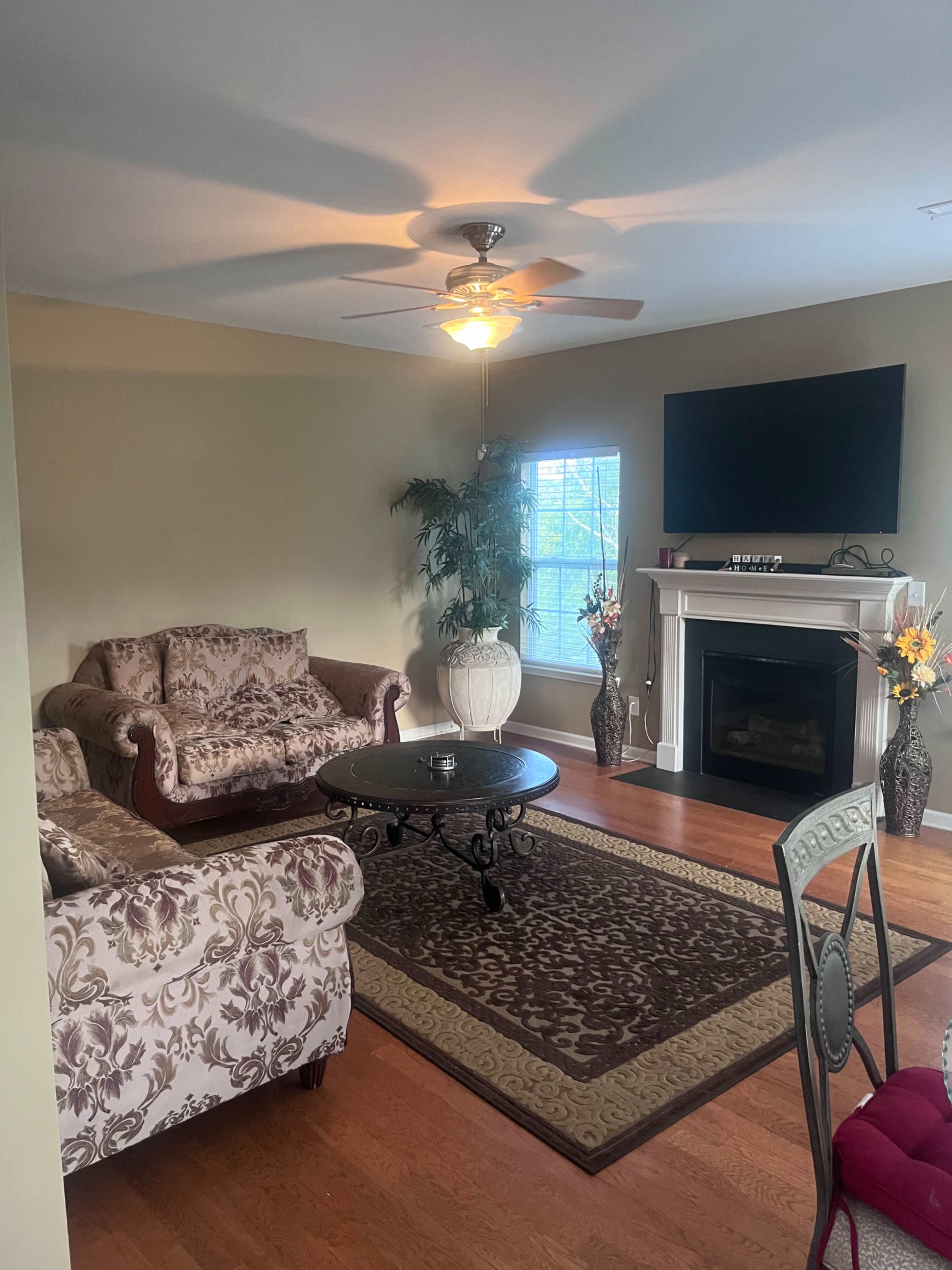 The living room features a floral-patterned couch and loveseat, a round coffee table, a fireplace with a television above it, and large windows allowing natural light to enter.