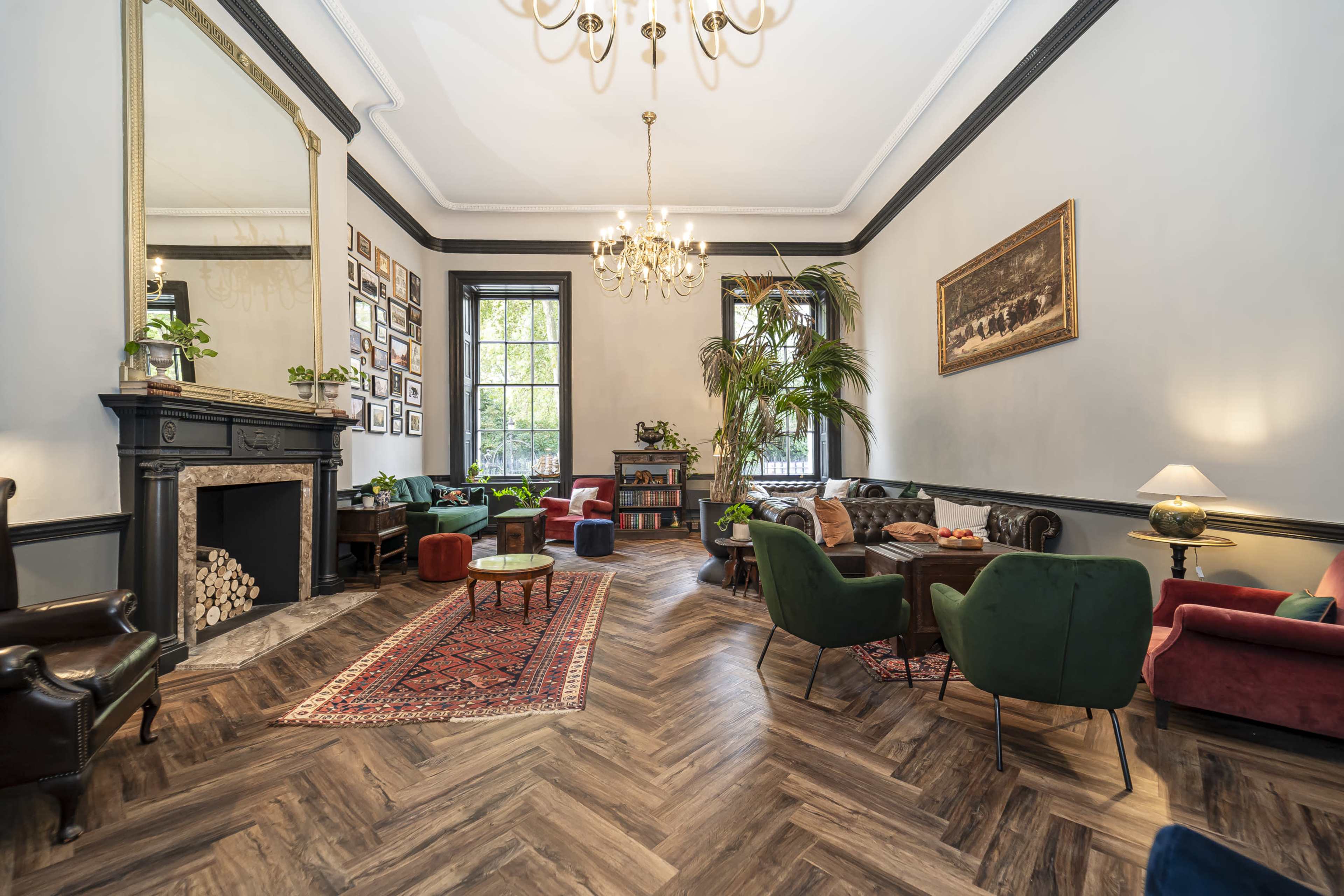 The image shows a stylish living room with a herringbone wood floor, a decorative fireplace, various seating arrangements, and large windows letting in natural light.