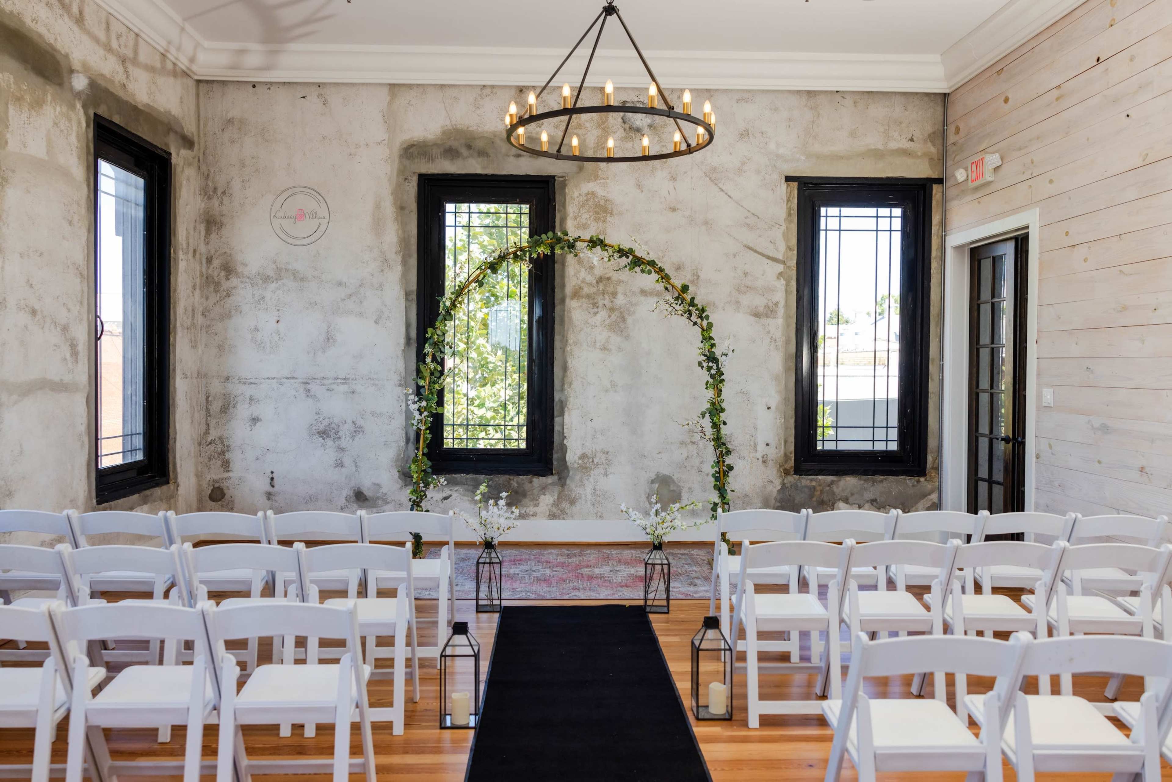 A simple wedding ceremony setup features white chairs arranged in rows facing a floral arch against a textured wall, with a black aisle runner leading to it.