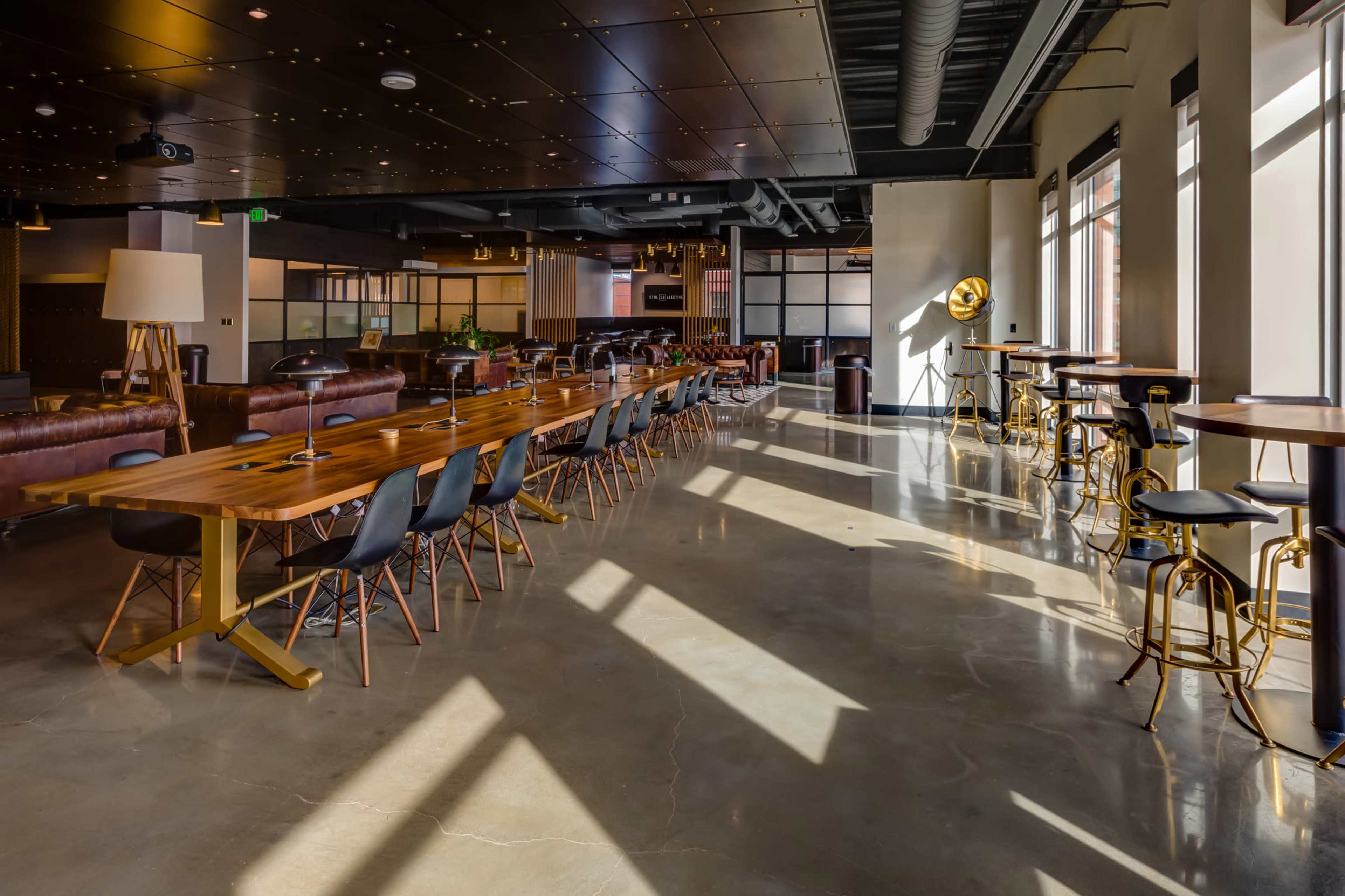 A modern open-concept workspace features a long wooden table, black chairs, and various seating areas illuminated by sunlight streaming through large windows.