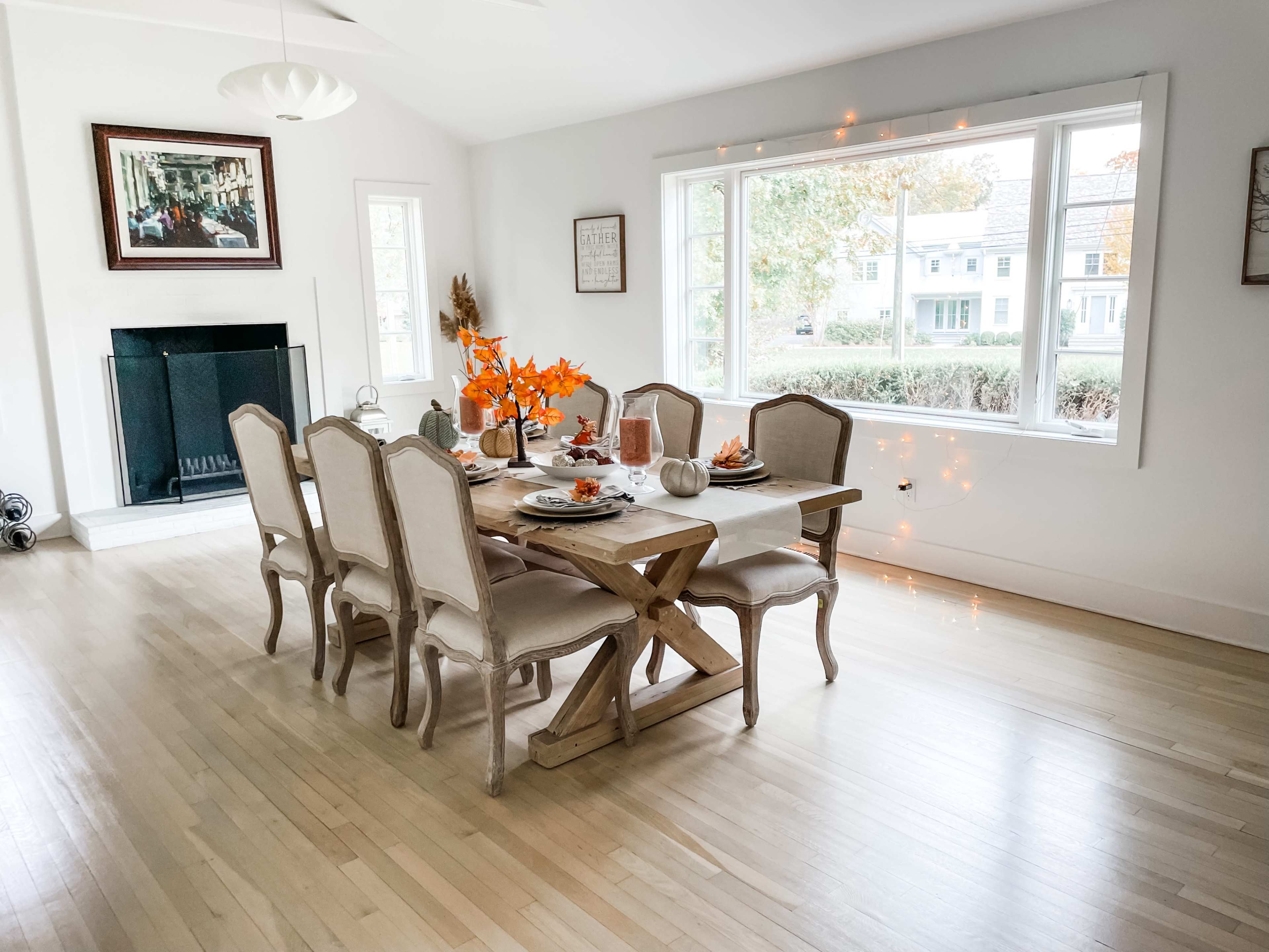 A dining room features a large wooden table set for a meal, surrounded by chairs, with orange autumn decorations and a view of a tree-lined street through the window.