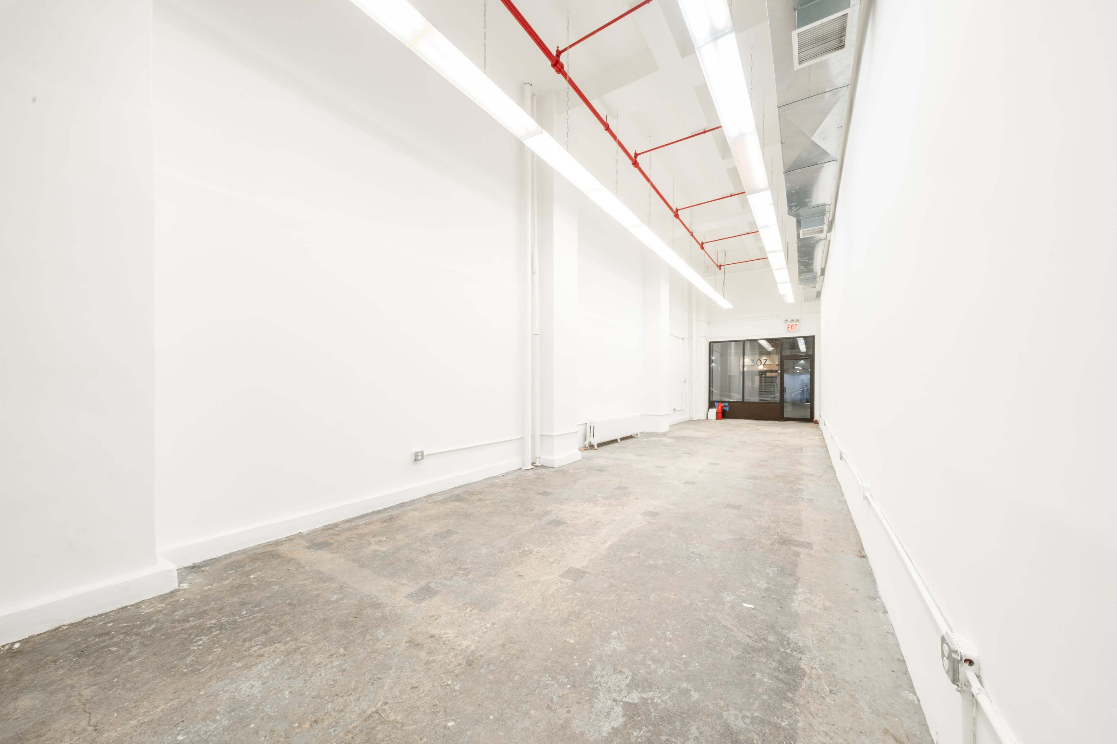 The image shows a spacious, empty room with white walls, a concrete floor, and large windows at one end.