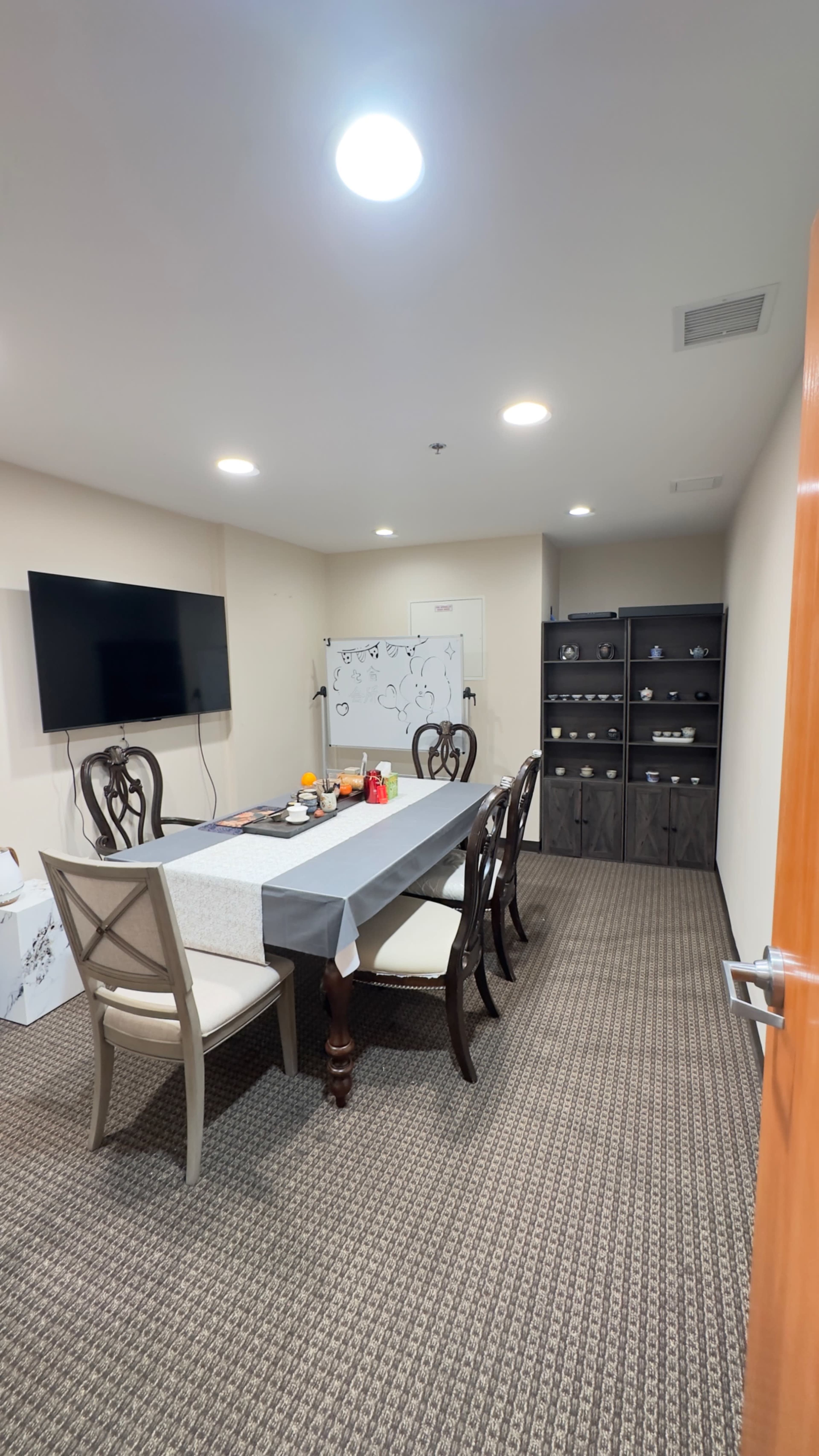 The image shows a conference room featuring a long table with chairs, a large wall-mounted television, a whiteboard, and a shelving unit filled with items.