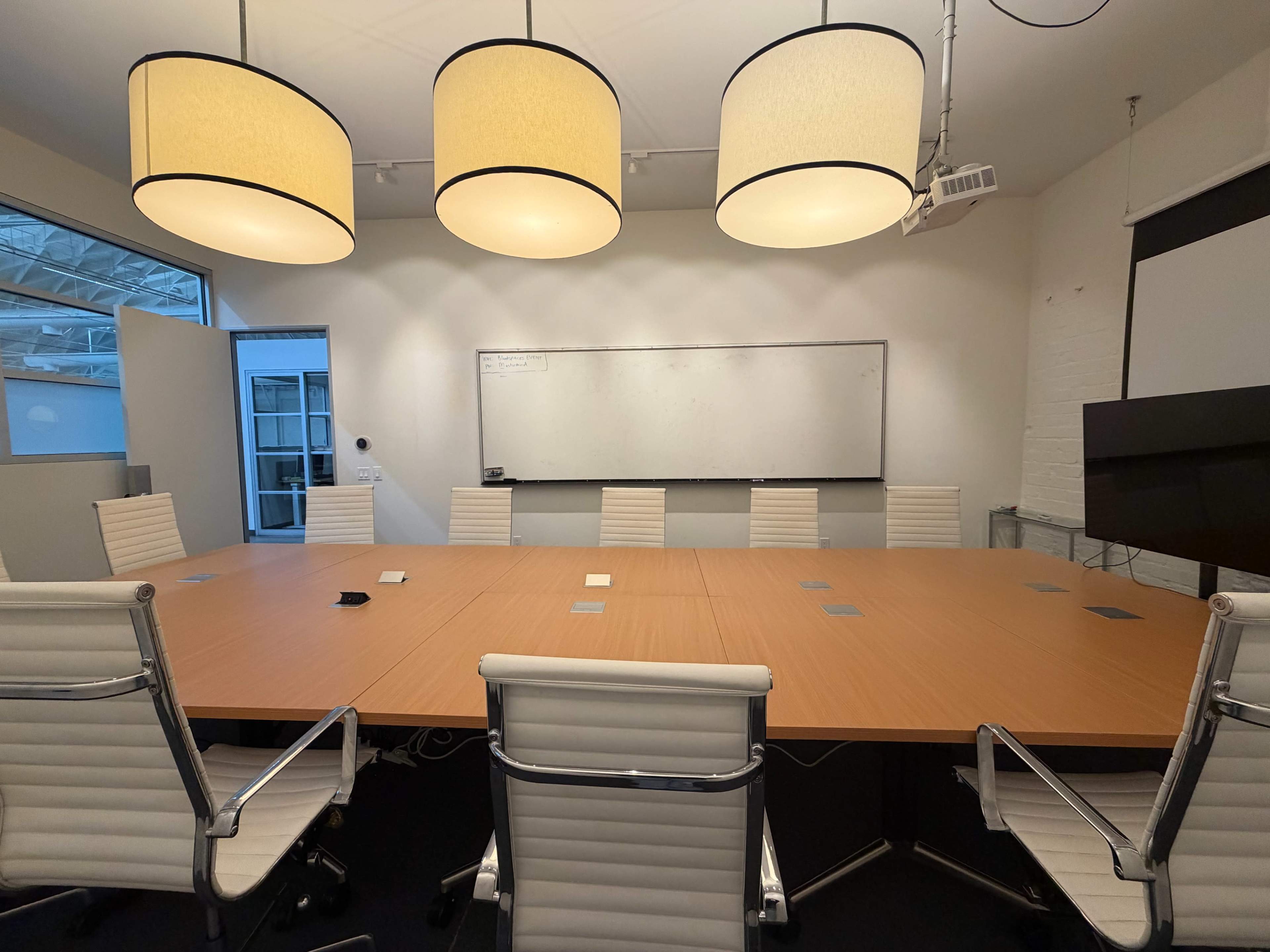 A large conference room features a long wooden table surrounded by white ergonomic chairs, with three pendant lights hanging above and a whiteboard on one wall.