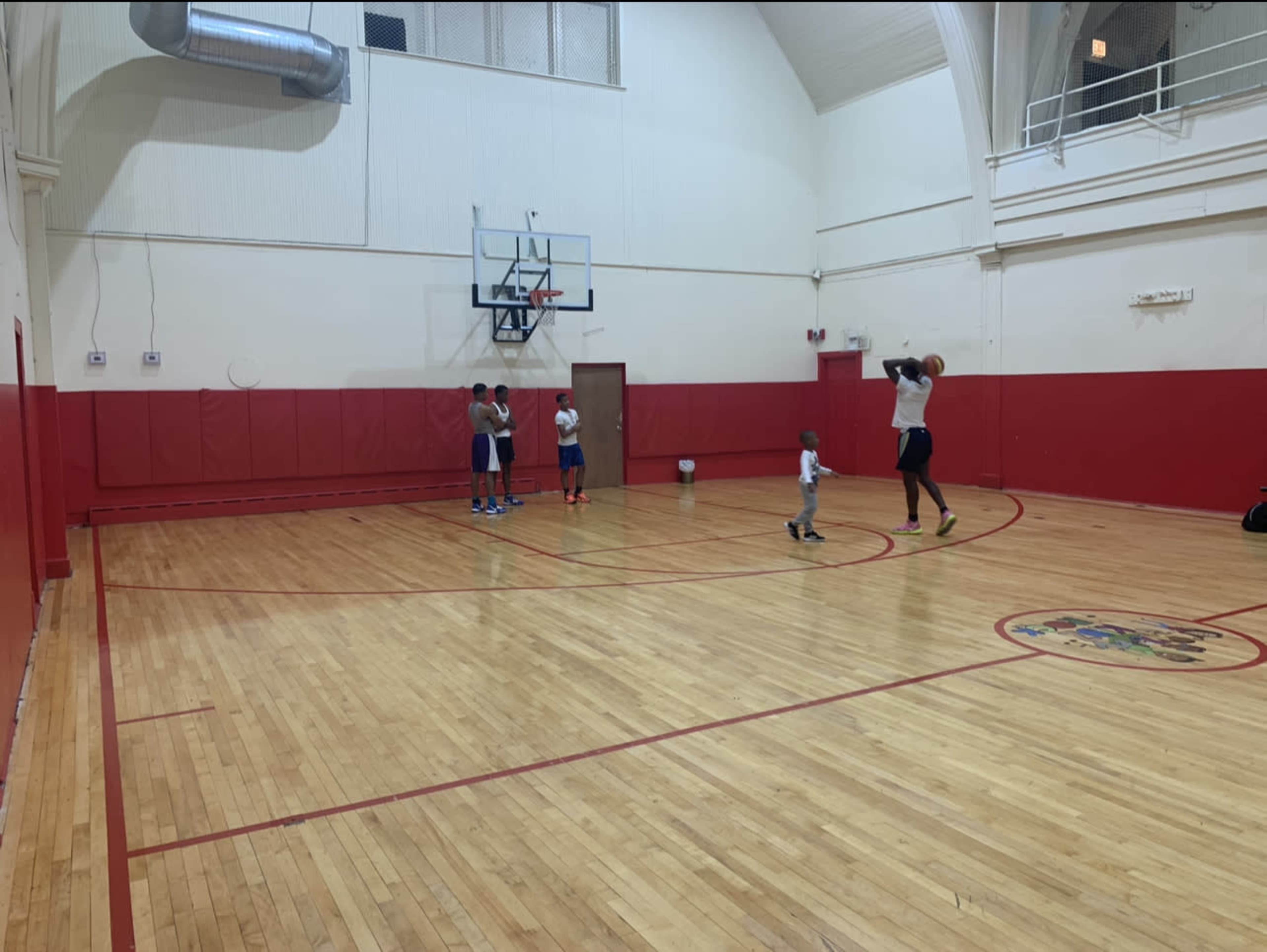A group of individuals practices basketball on a hardwood gym floor with red walls and a hoop at one end.
